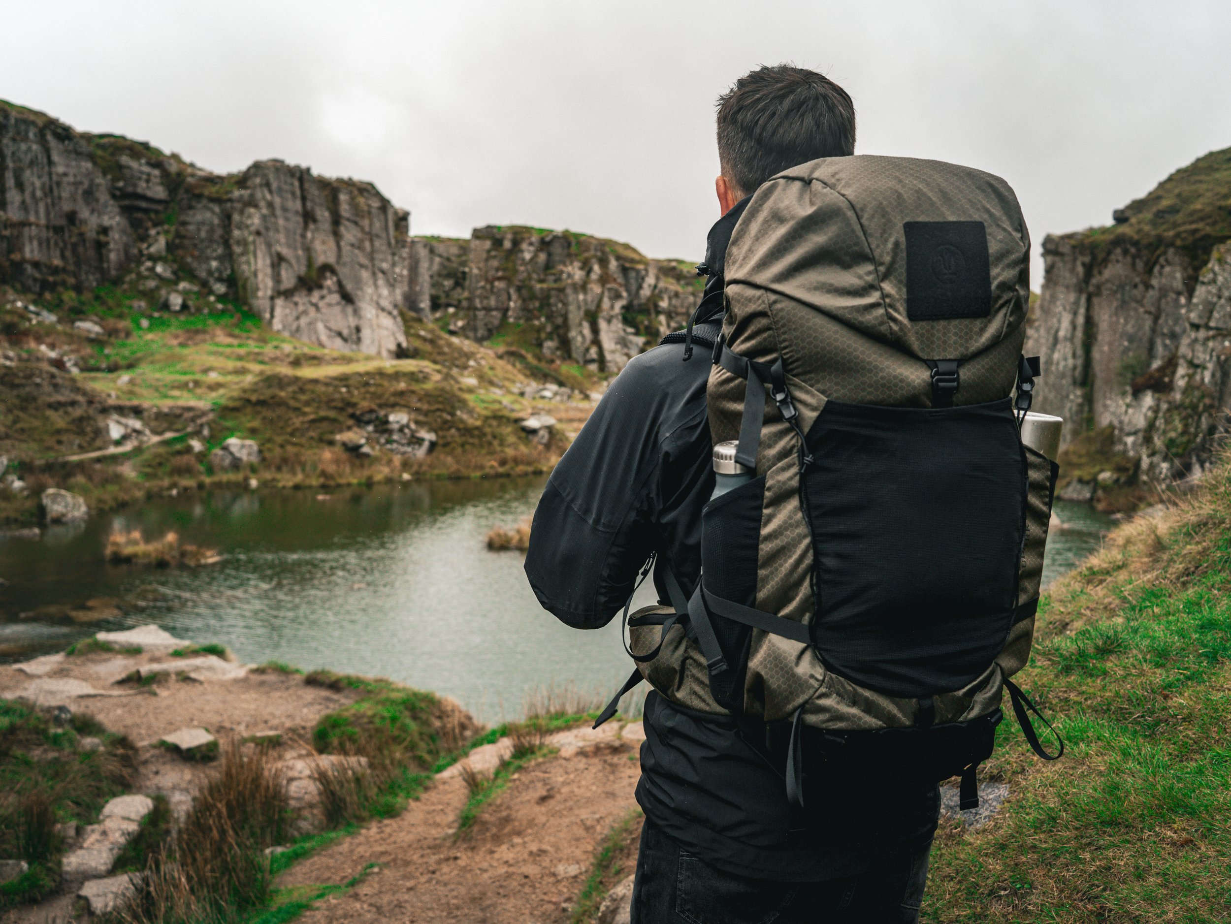 A person with short hair and a beige backpack sitting on a rock, overlooking a body of water with rocky hills in the background on a cloudy day.