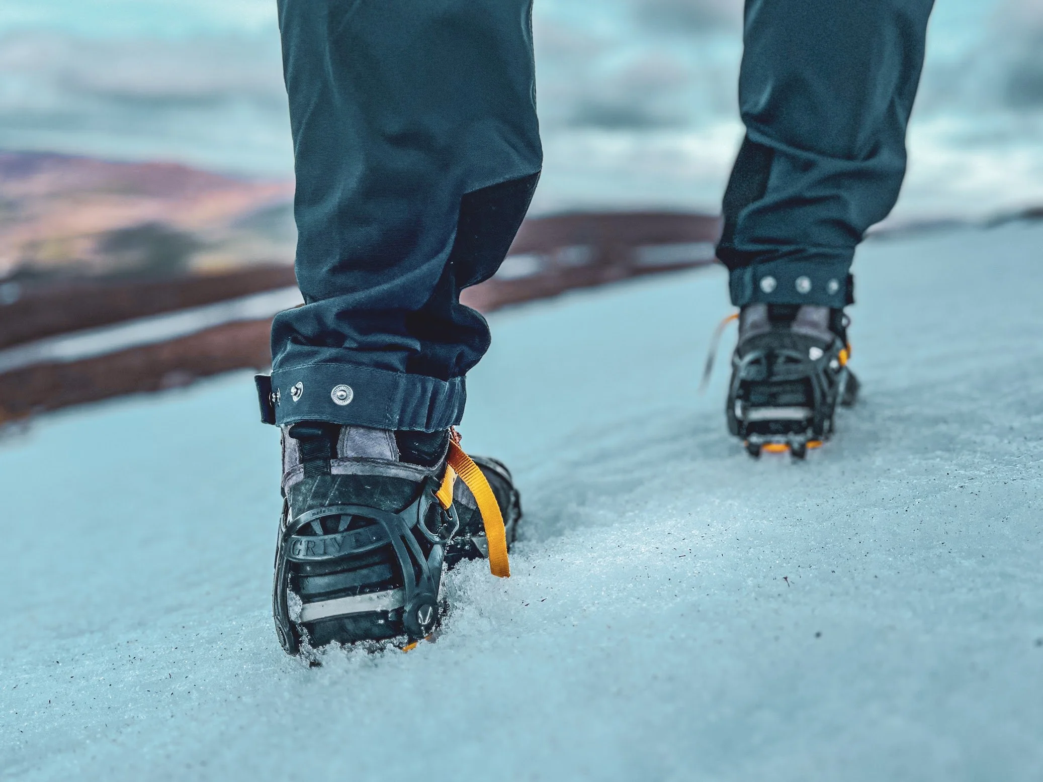 Close-up of a person wearing black winter boots and blue jeans walking on snow-covered ground with a shadow of a bench in the background.