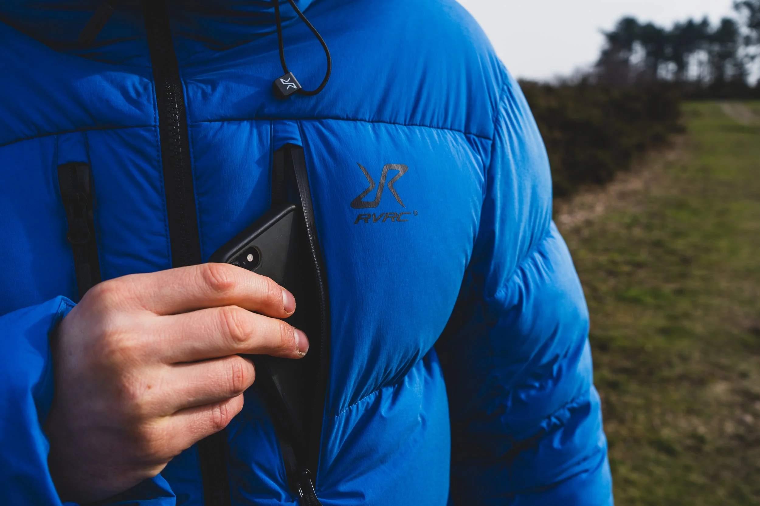 A person wearing a blue outdoor jacket with a logo on the chest, holding a smartphone and wearing Bluetooth earphones, outdoors with trees and grass in the background.