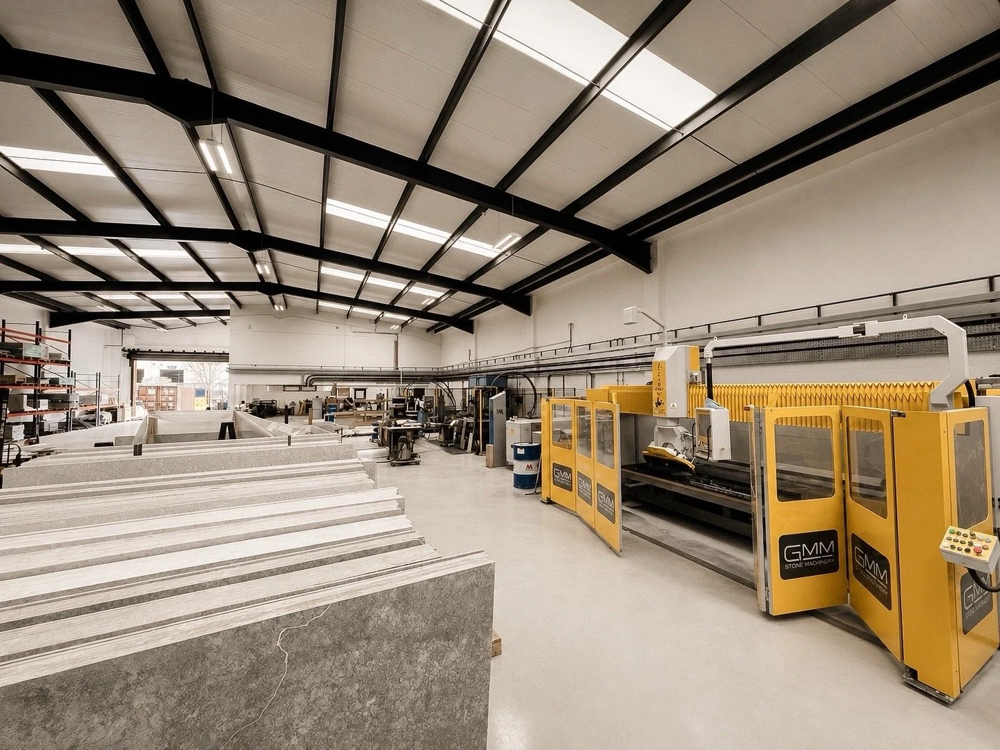 Inside a stone manufacturing shop with large machinery, work tables, and shelves filled with materials, under a high ceiling with black beams and fluorescent lighting.