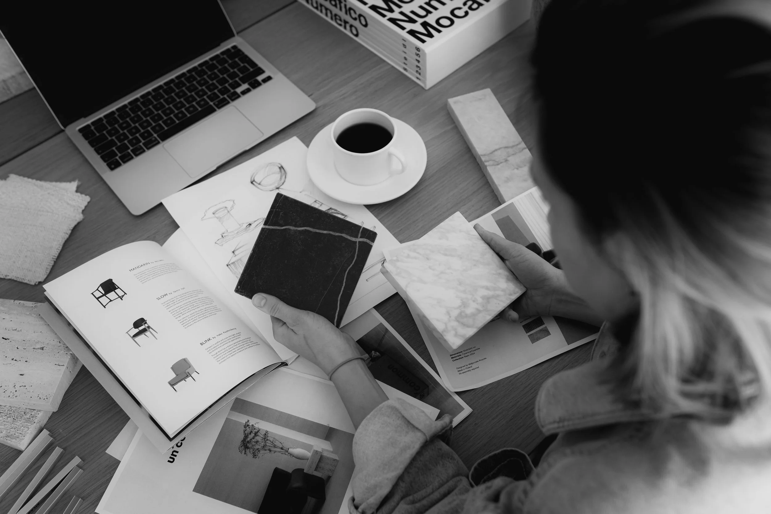 A person is looking at samples of marble and tile swatches on a table, with books and a laptop open nearby, possibly working on interior design ideas.