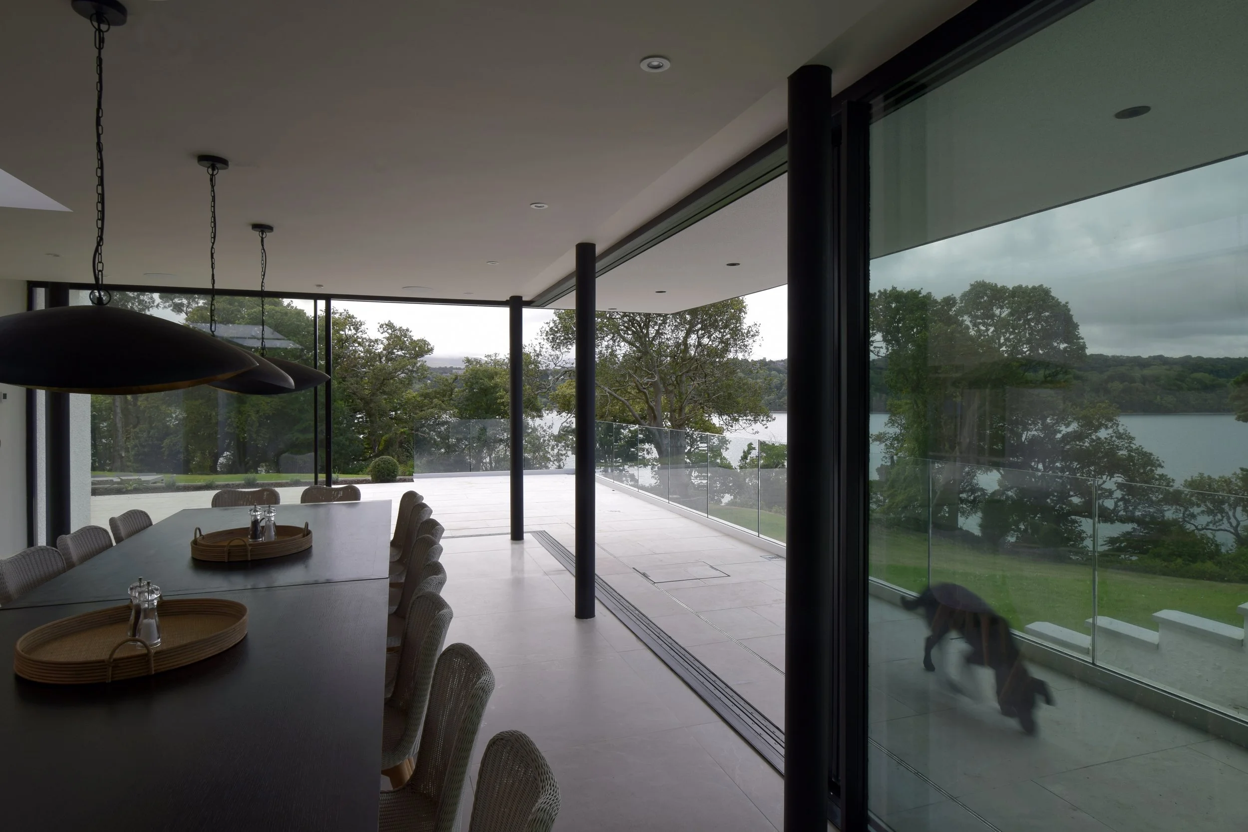 Modern dining area with a long table, chairs, and large glass windows overlooking a lake and trees. A dog is outside on the patio.