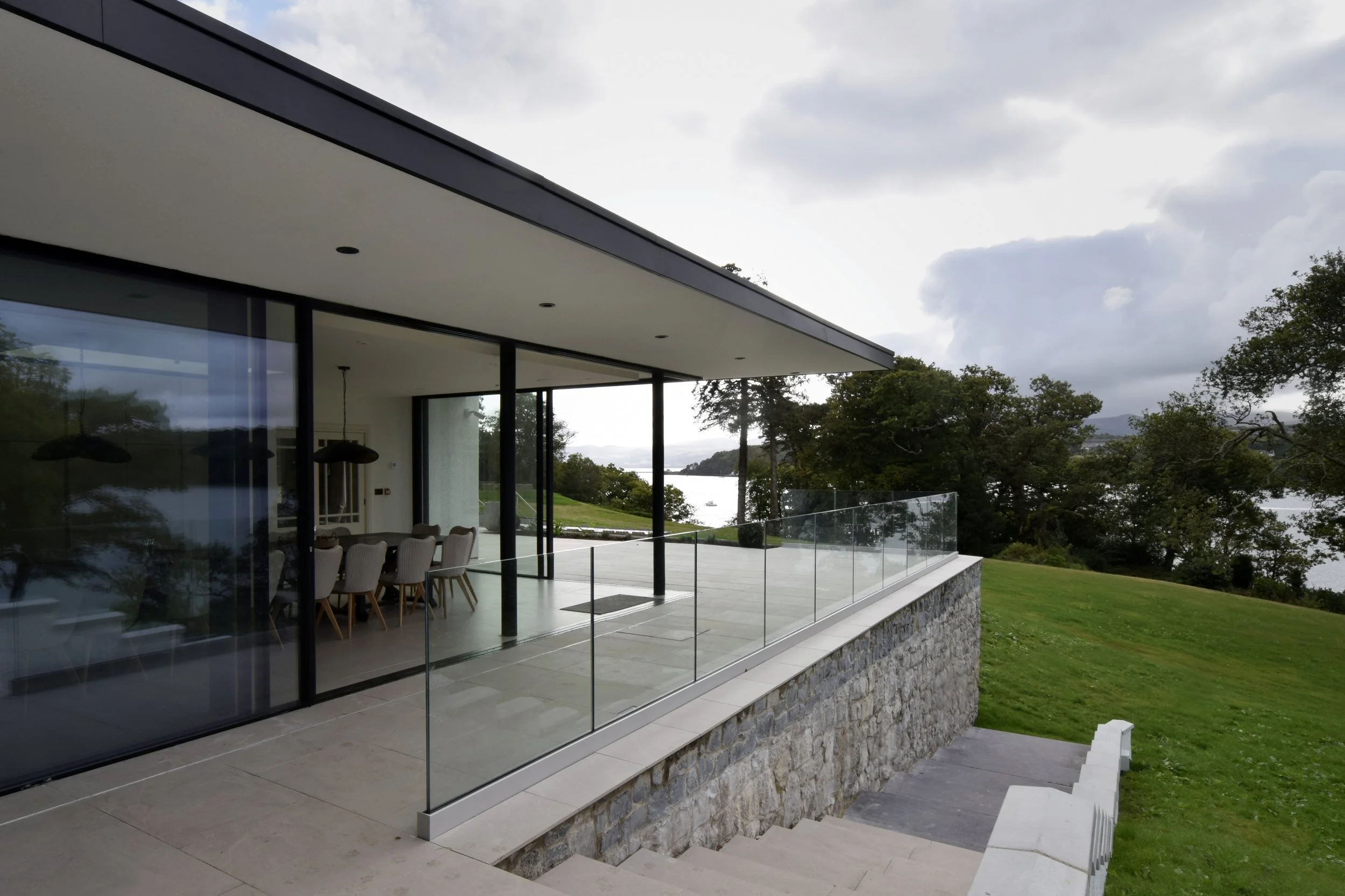Modern house with large glass sliding doors and a glass railing, overlooking a lush green lawn, trees, and a body of water in the distance, under a partly cloudy sky.