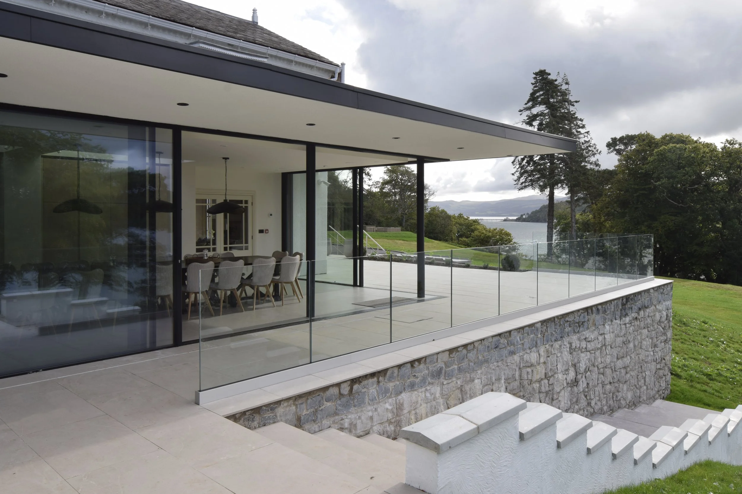 Modern house with large glass windows and a spacious balcony overlooking a body of water, surrounded by trees and green landscape.