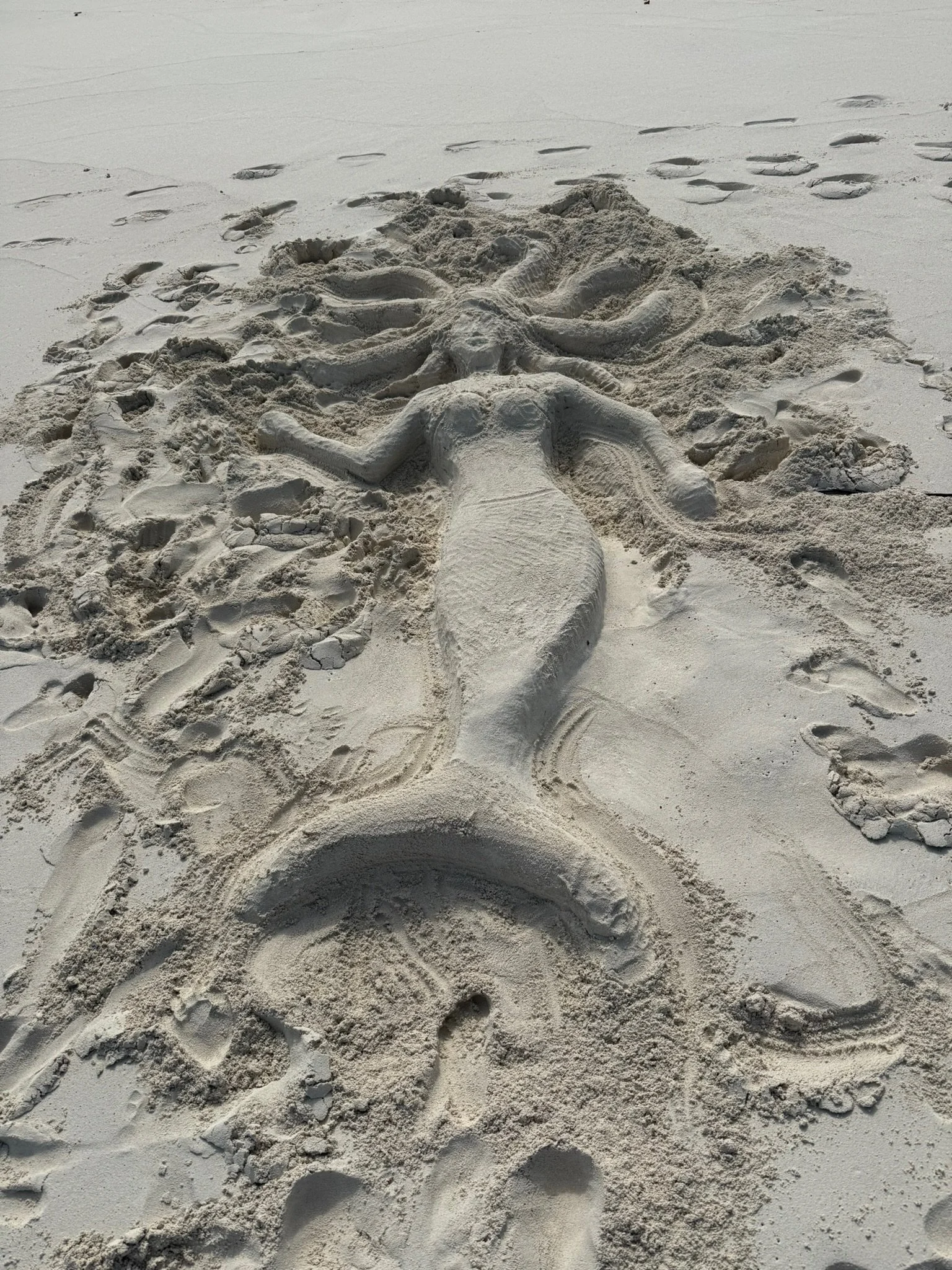 Sand sculpture of a mermaid with flowing hair and a fish tail, lying on the beach with footprints around.