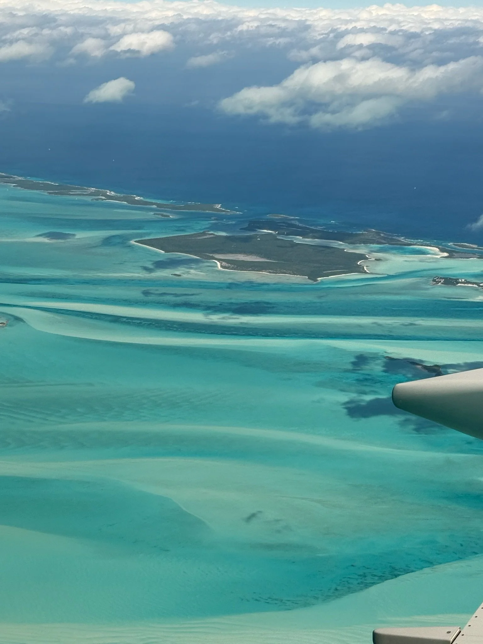 Aerial view of turquoise and blue ocean water with sandbars and small islands, under a partly cloudy sky.