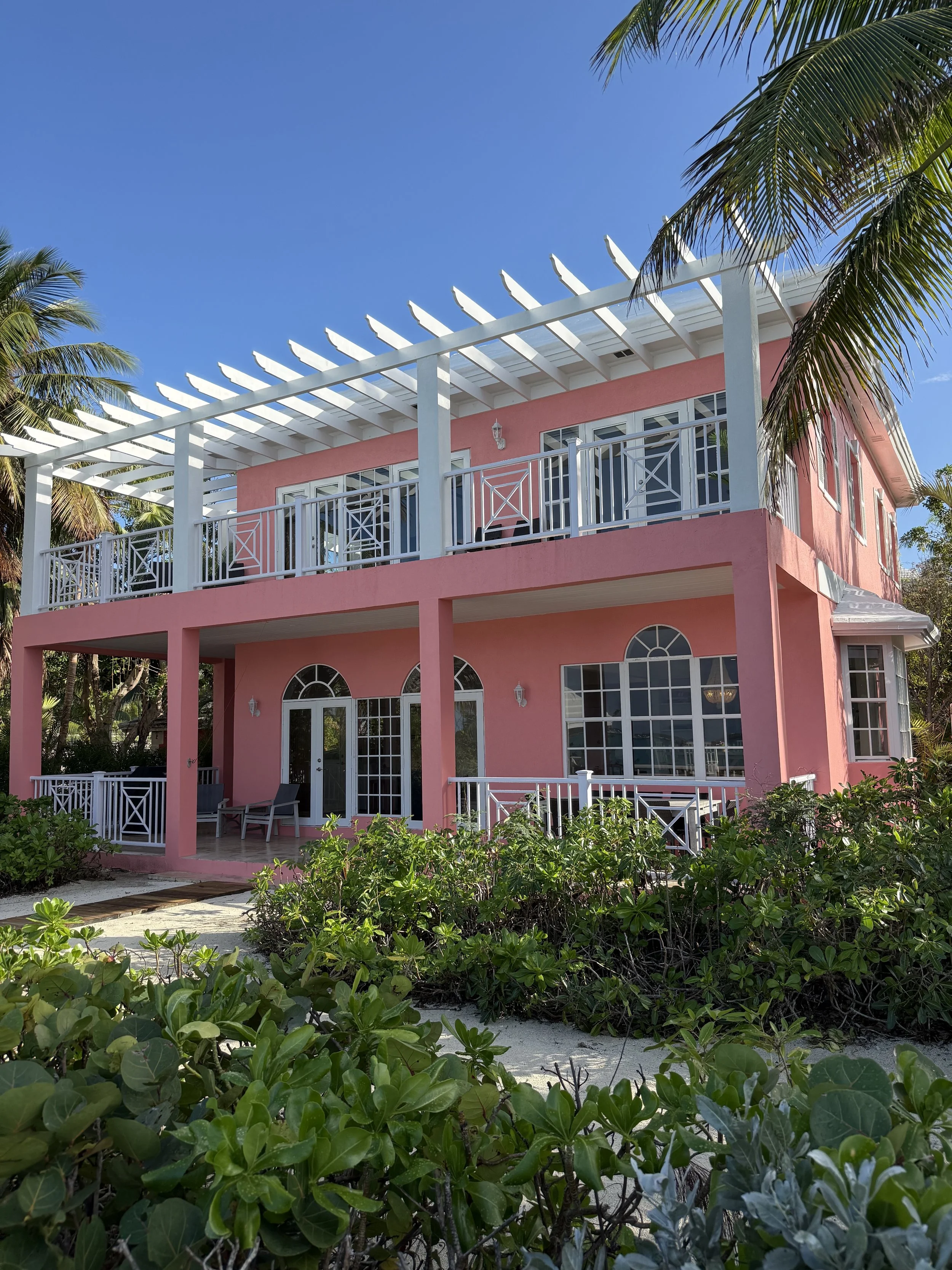 A pink two-story house with white railings and a white pergola on the upper balcony, surrounded by green bushes and palm trees under a clear blue sky.