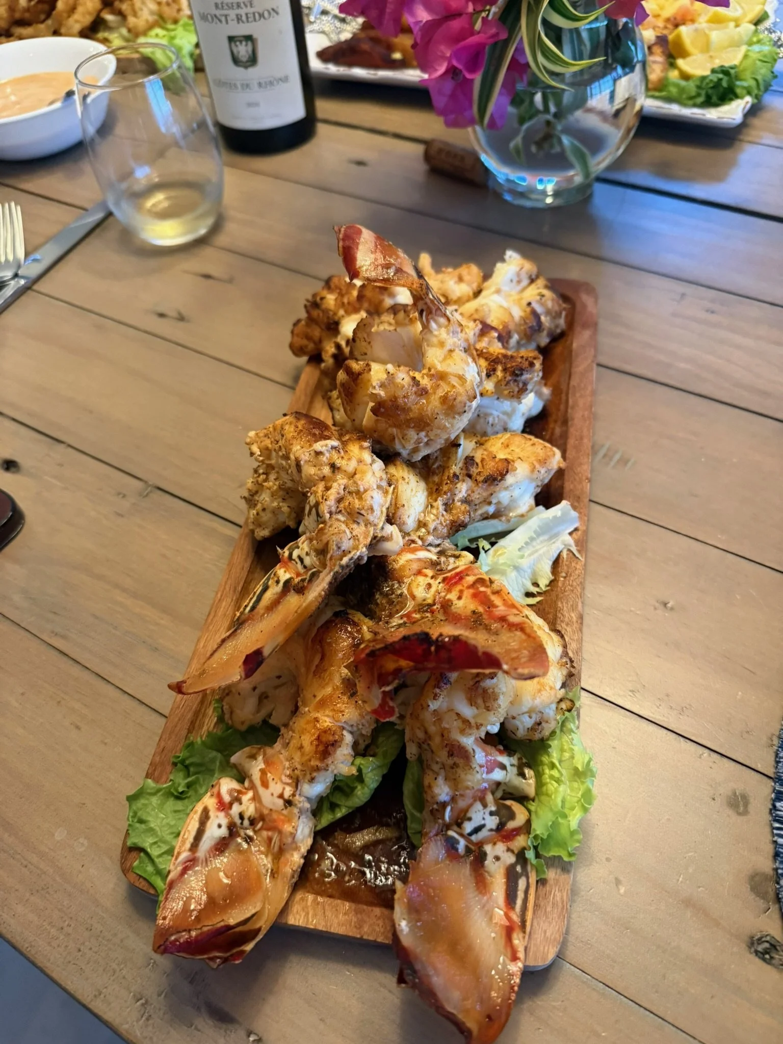 A wooden serving board with cooked lobster tails, garnished with green lettuce, on a wooden table. Surrounding items include a wine bottle, a glass of white wine, a bread roll, a bowl with dipping sauce, and a vase with pink and white flowers.