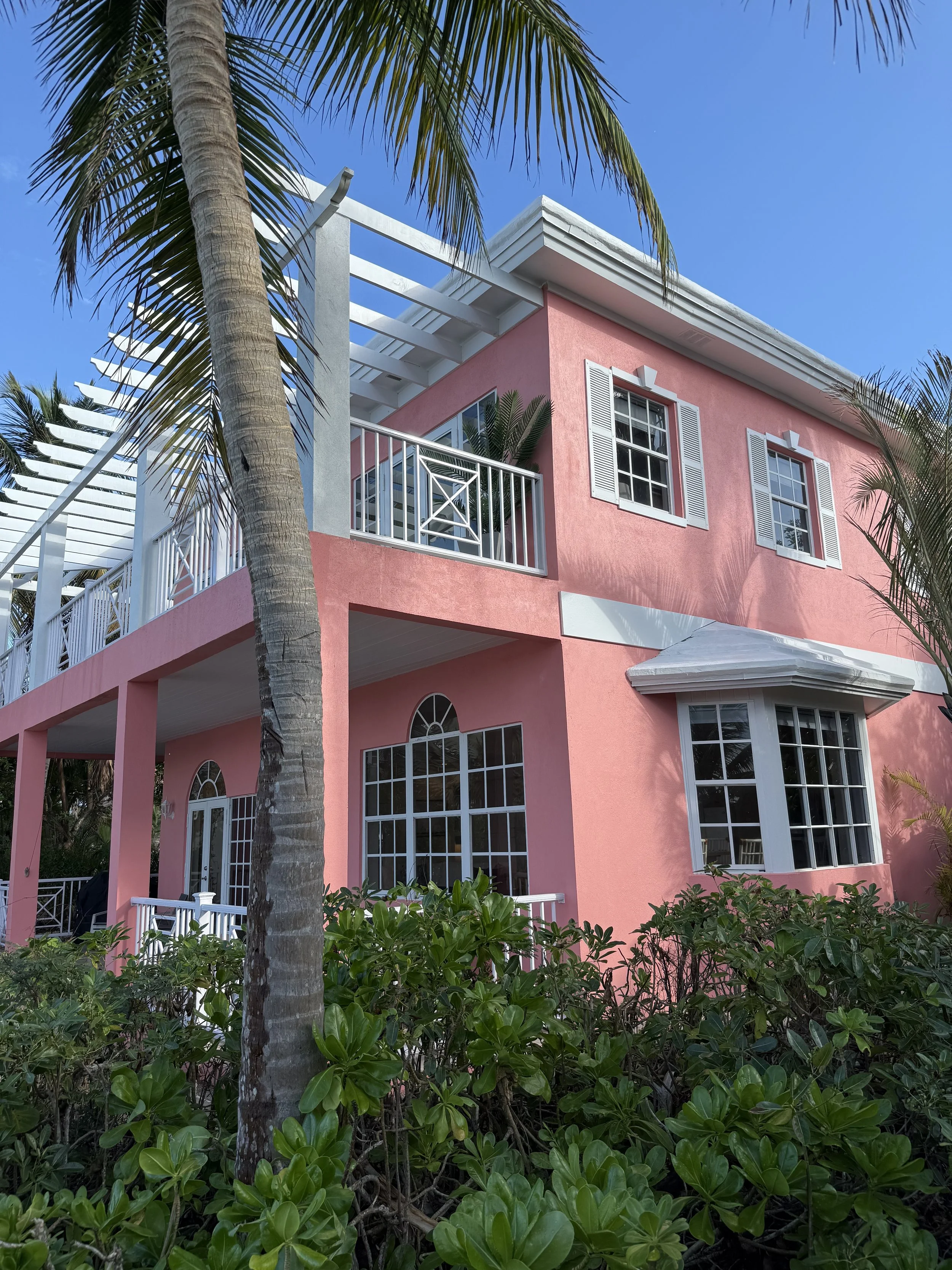 Pink multi-story house with white shutters, large windows, and a white balcony, surrounded by green plants and palm trees, under a clear blue sky.