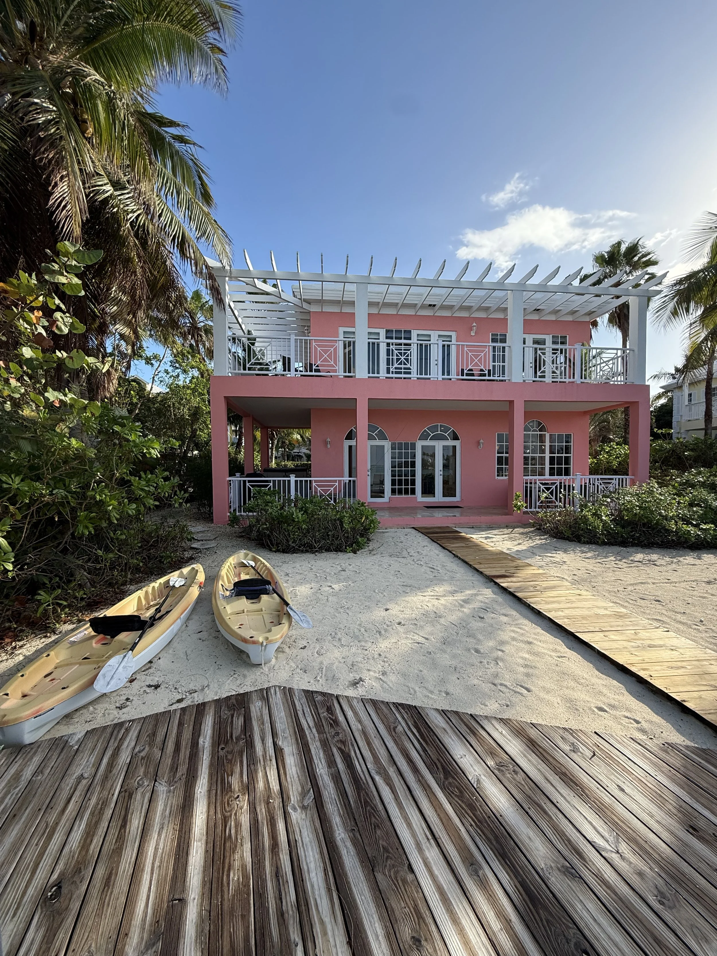 A two-story pink beach house with white railings and a covered balcony on each level, surrounded by palm trees, with sandy grounds, two kayaks with paddles on the sand, a wooden walkway, and a bright blue sky with a few clouds.