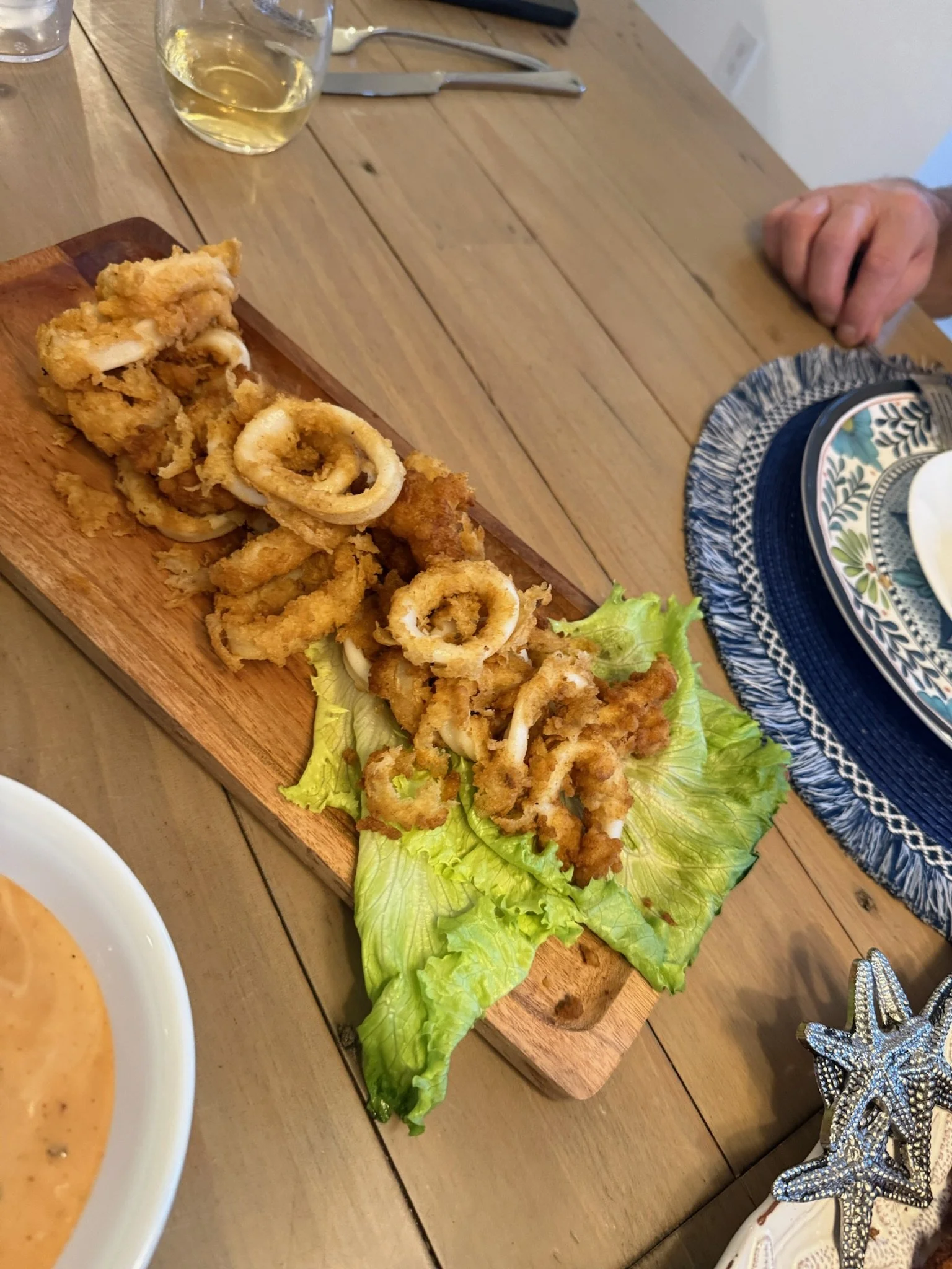 Fried calamari served on a wooden board with lettuce, accompanied by a dipping sauce, on a wooden table with a wine glass and cutlery.