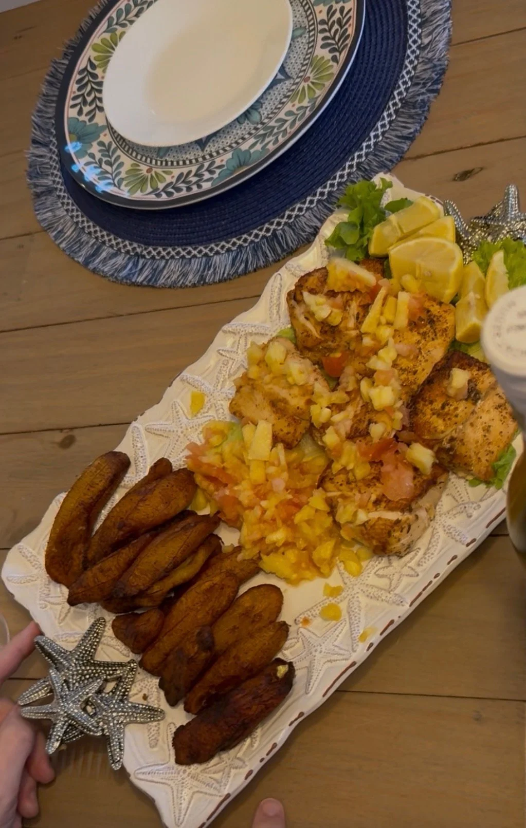 A dinner plate stand with grilled fish topped with pineapple and onions, a side of fried plantains, lemon wedges, and lettuce garnishes on a white decorative tray.