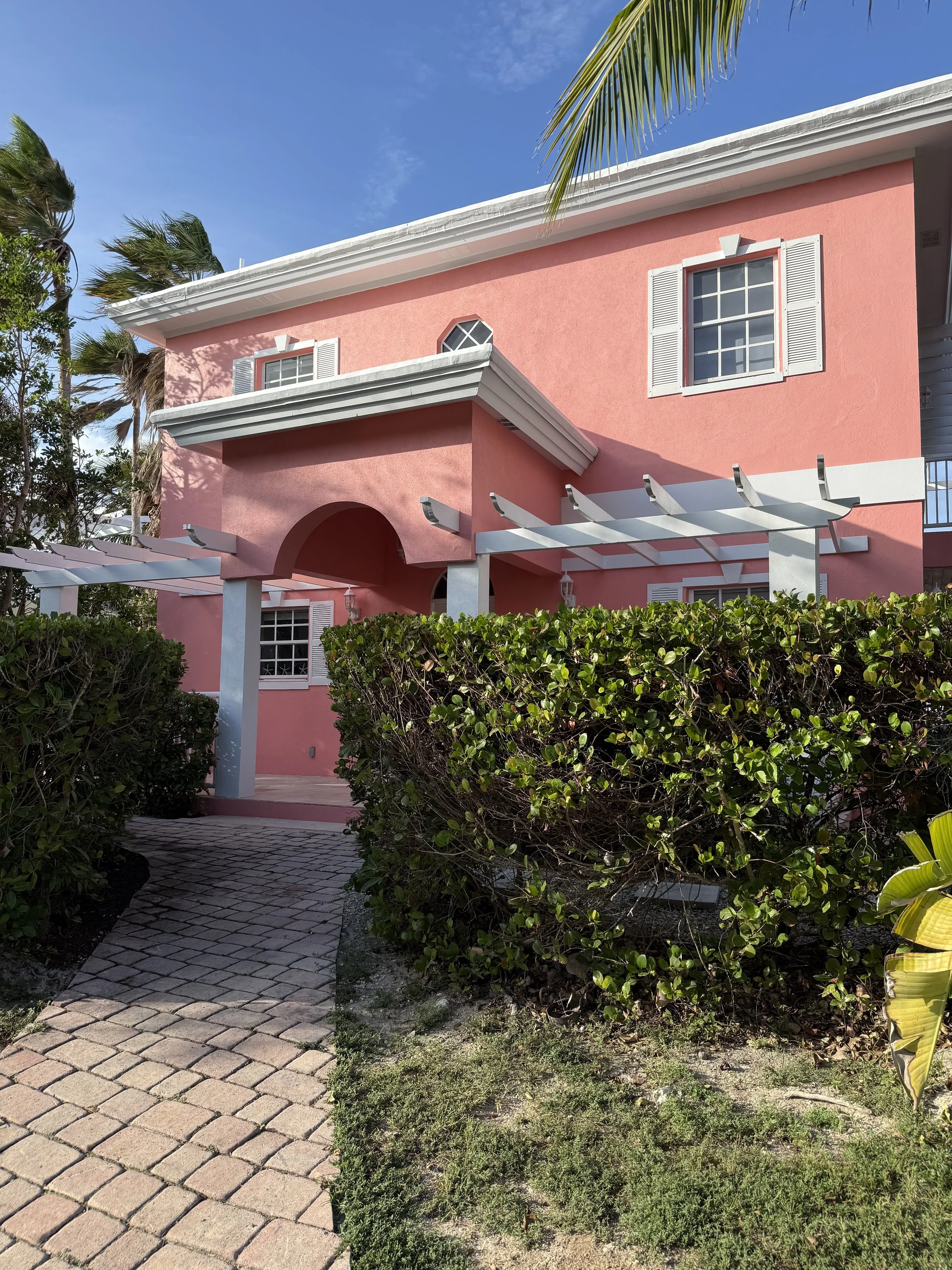 A pink two-story house with white window shutters and a white pergola in front, surrounded by greenery, under a partly cloudy blue sky.
