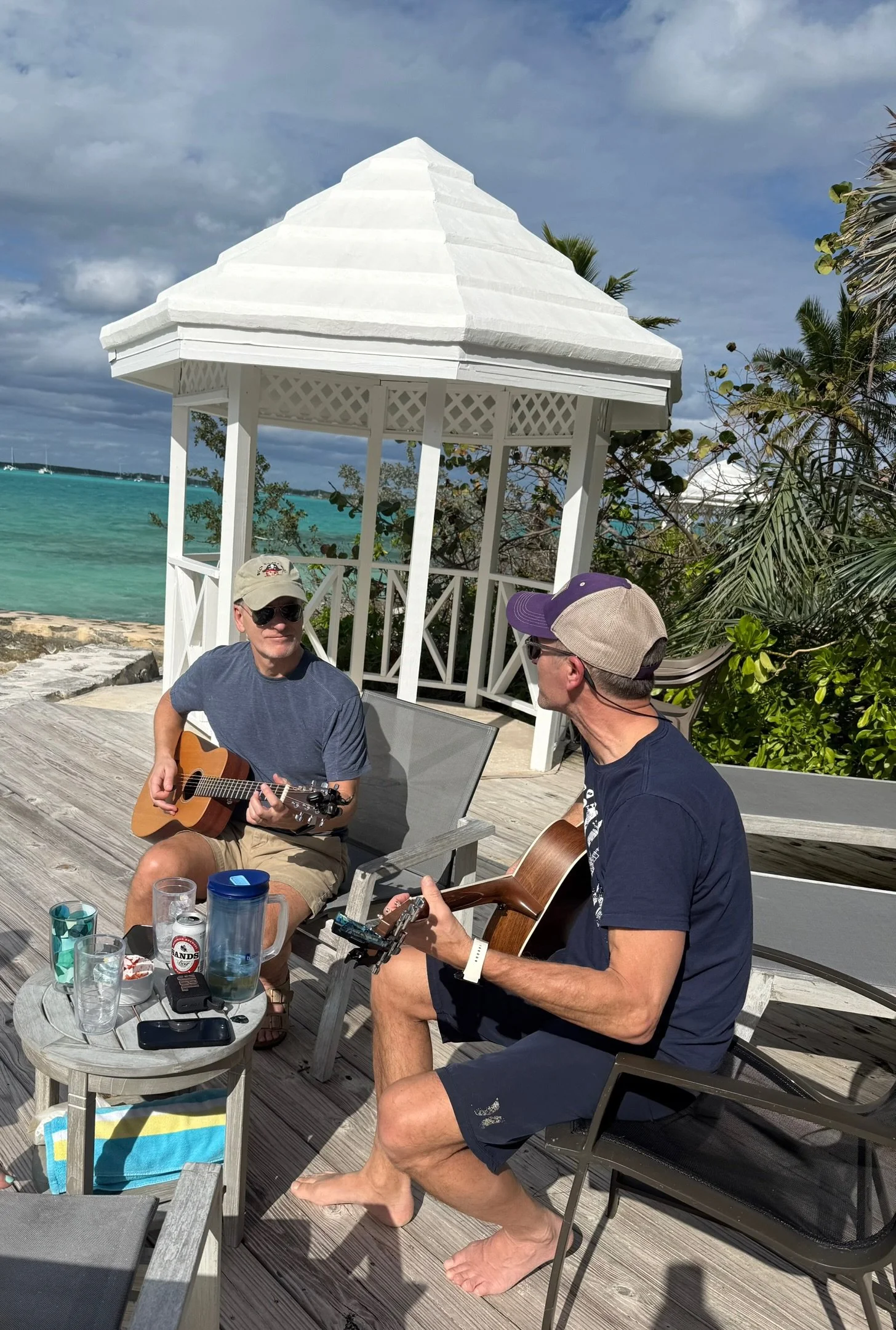 Two men playing acoustic guitar and singing on a wooden deck near the ocean. One is wearing sunglasses, a beige cap, and a navy shirt, while the other is wearing a beige cap, sunglasses, and a navy shirt. There is a small table with drinks and a towe