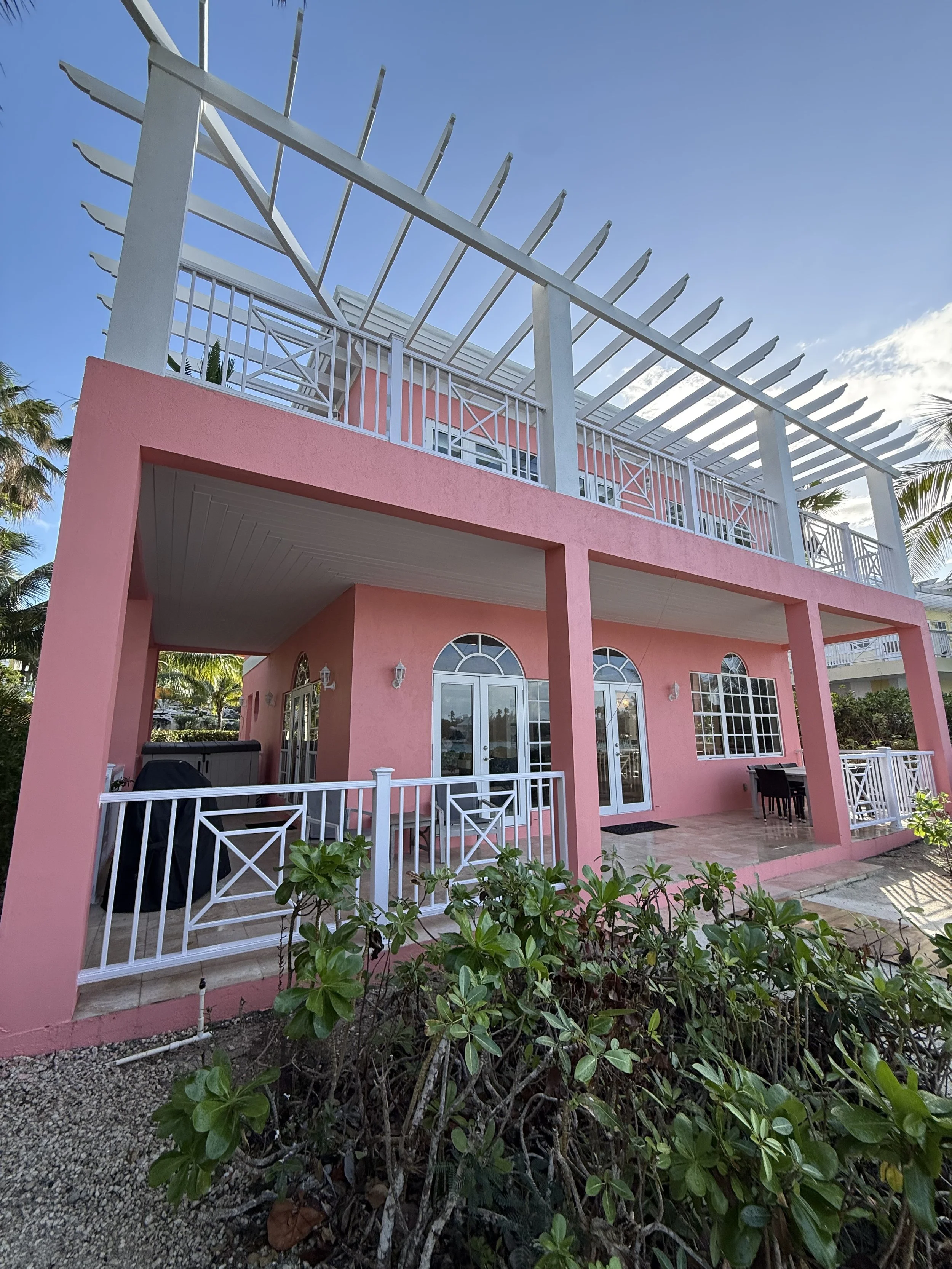 A two-story house painted in pink with white trim, featuring a balcony on the upper level with white railings and a pergola, surrounded by palm trees and greenery under a clear blue sky.