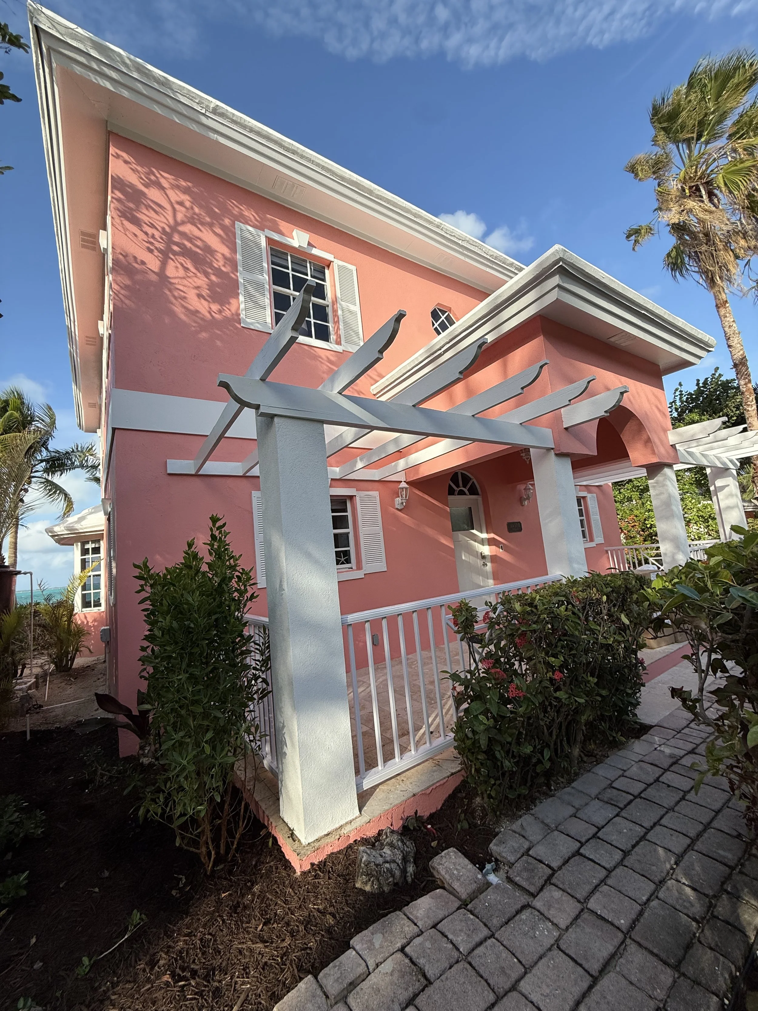 Pink house with white trim, white pergola, and a pebble pathway with bushes in front. Palm trees and blue sky in the background.