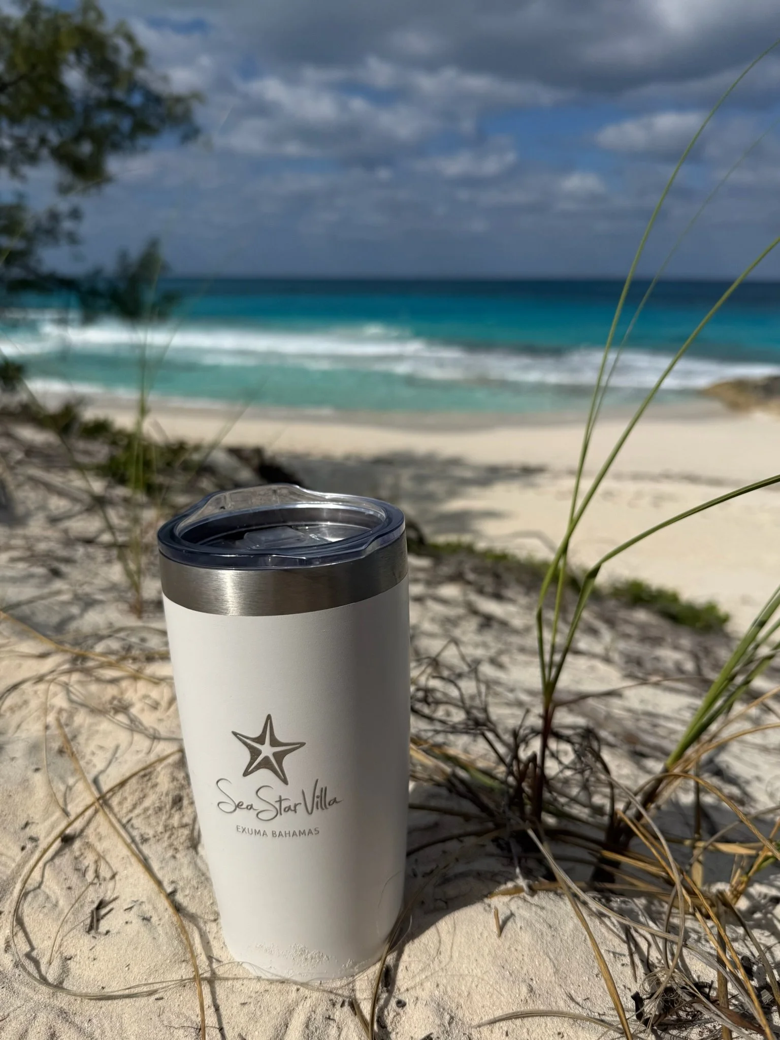 A white insulated tumbler with a starfish logo and the words 'Sea Star Villa EXUMA BAHAMAS' sits on sandy beach dune grass, with turquoise ocean waves and a partly cloudy sky in the background.