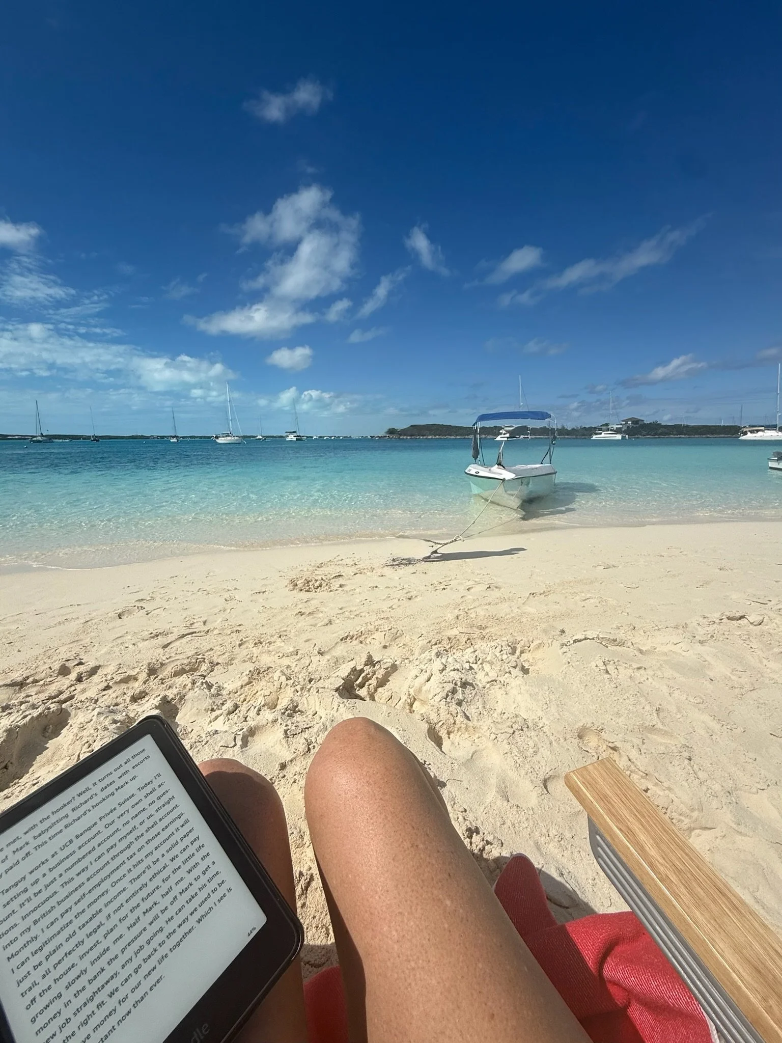 A person relaxing on a sandy beach, reading on a tablet, with a boat anchored nearby and more boats sailing in the clear blue ocean under a partly cloudy sky.