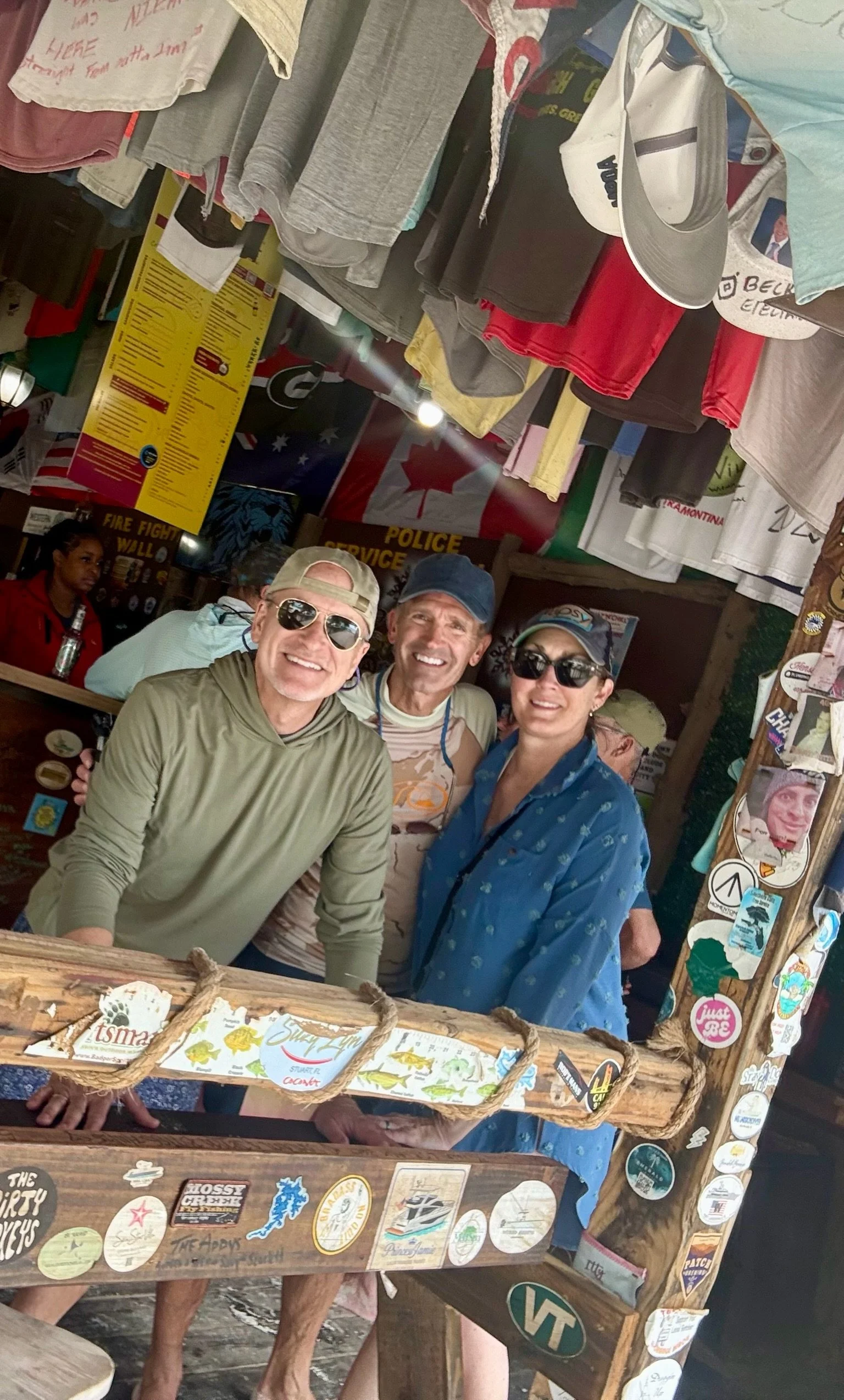 Three people smiling, standing under hanging clothes and surrounded by stickers and signs in a casual outdoor setting.