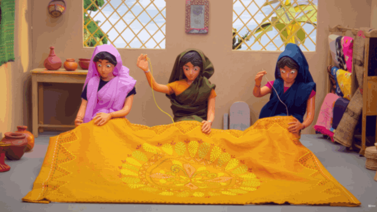 Three women sewing a large yellow embroidered textile together inside a workshop.