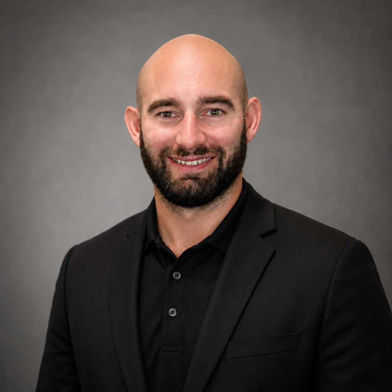 A man with a beard and shaved head smiling, wearing a black t-shirt with 'millennium family fitness' logo, against a black background.