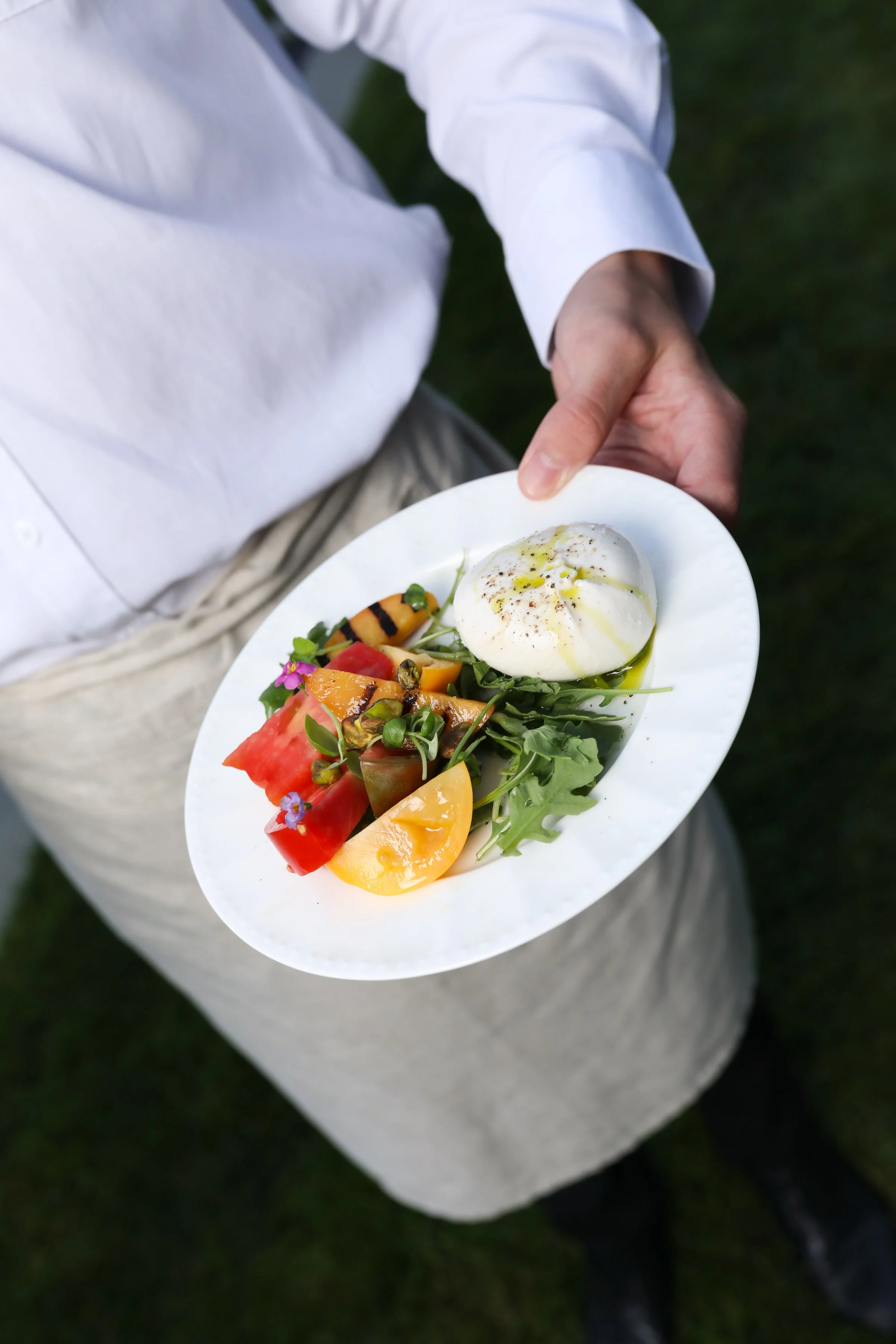 A person in a white shirt holding a white plate with a caprese salad featuring sliced tomatoes, fresh herbs, a mozzarella ball drizzled with olive oil, and a garnish of microgreens against a dark green grassy background.