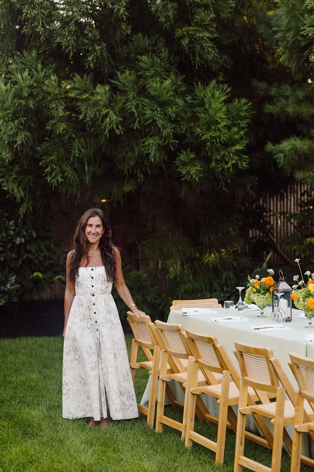 A woman in a white, floral dress stands outdoors next to a long dining table decorated with flowers and tableware, with large green trees in the background.