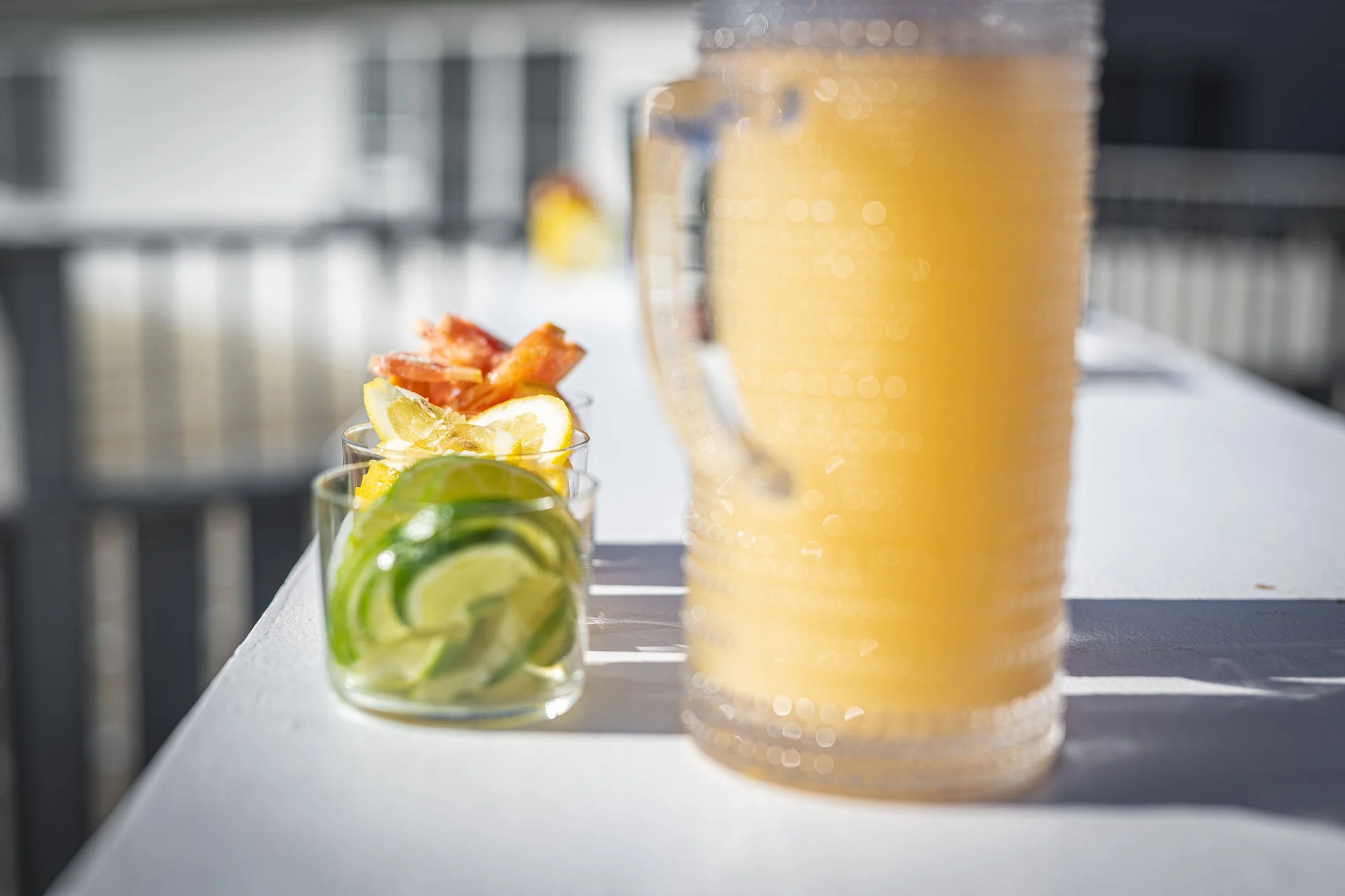 A row of beverages on a white surface, including a glass of sliced lemon, a slice of grapefruit, and slices of lime, with a blurred background of bottles and a glass of beer.