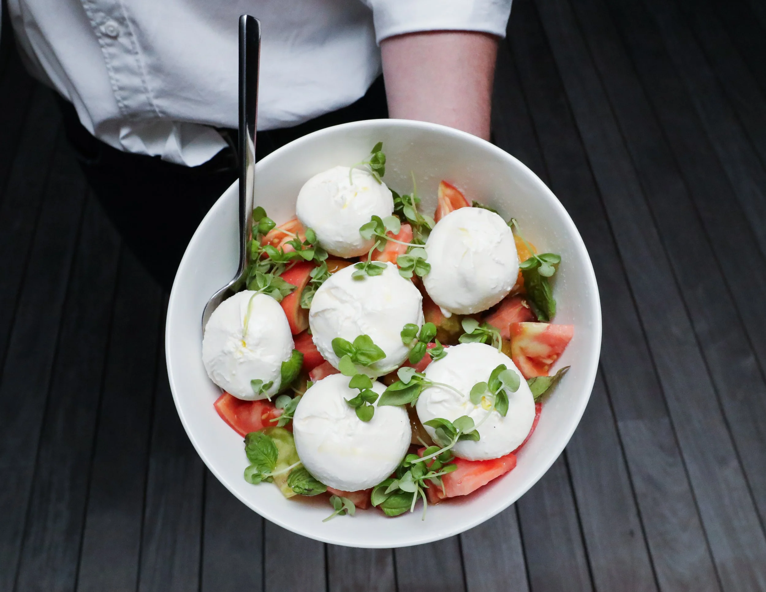 A white bowl filled with sliced tomatoes, fresh microgreens, and small mozzarella balls on a dark wooden table.