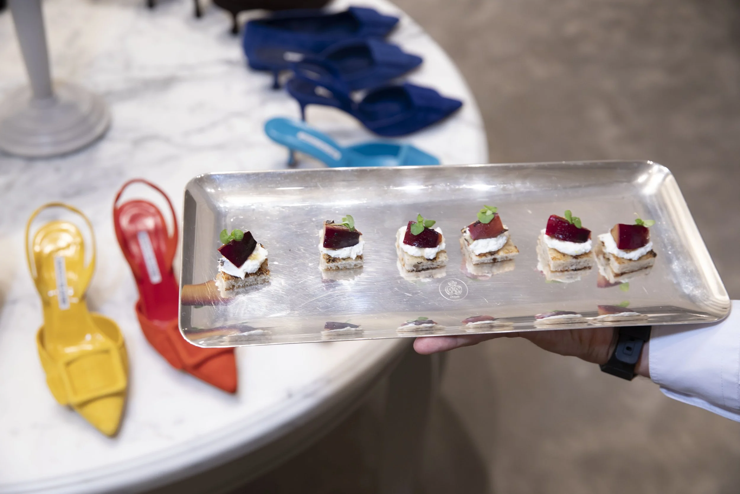 Small appetizer bites on a silver tray, topped with cream, red jelly, and microgreens, held by a person wearing a white shirt and black watch. In the background, there are blue shoes, yellow and red items, and what appears to be a white tablecloth.