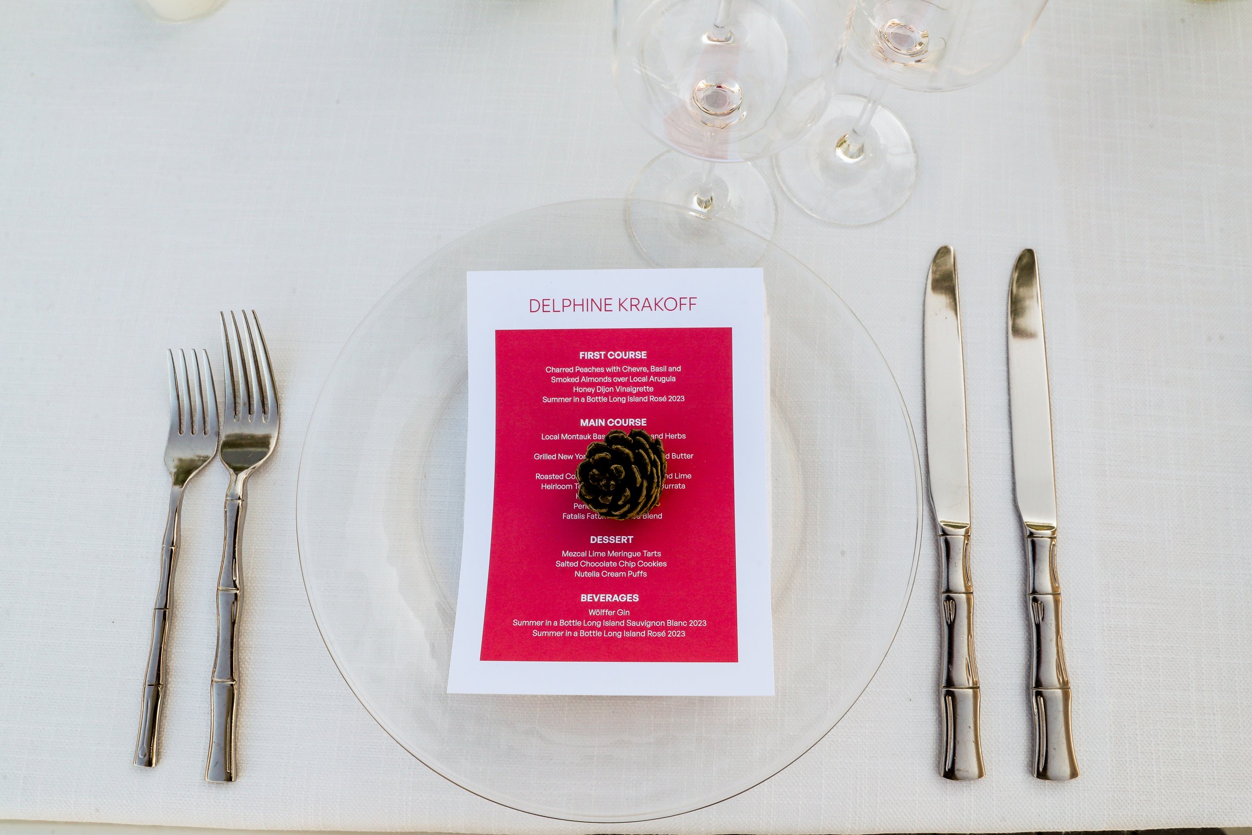 Table setting with two forks on the left, two knives on the right, a clear glass plate, a red and white printed menu, and three empty wine glasses above the menu, all on a white tablecloth.