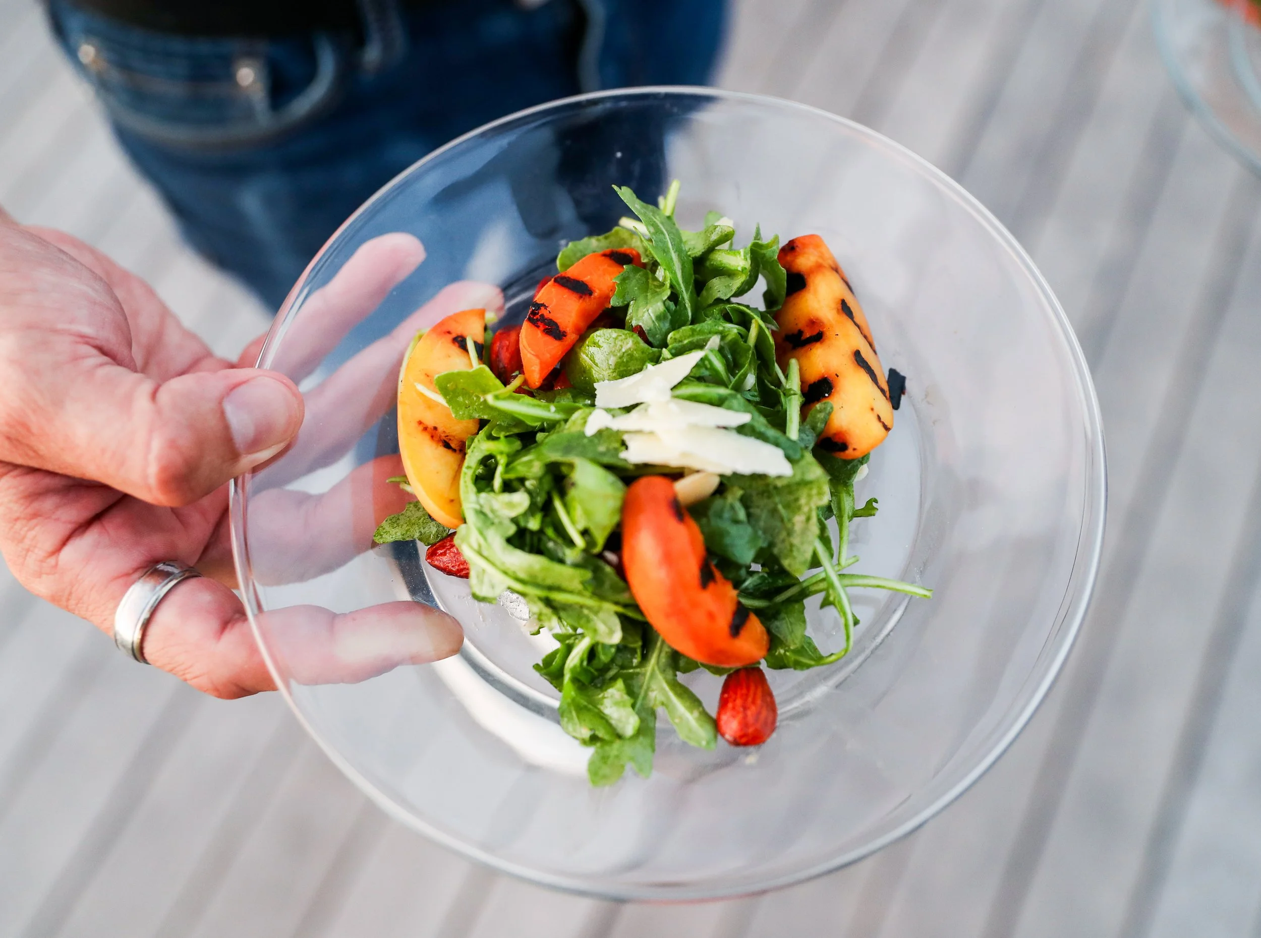 A clear glass bowl of mixed salad with leafy greens, grilled orange peppers, and shredded cheese, held by a person with a wedding ring, standing on a gray textured surface.