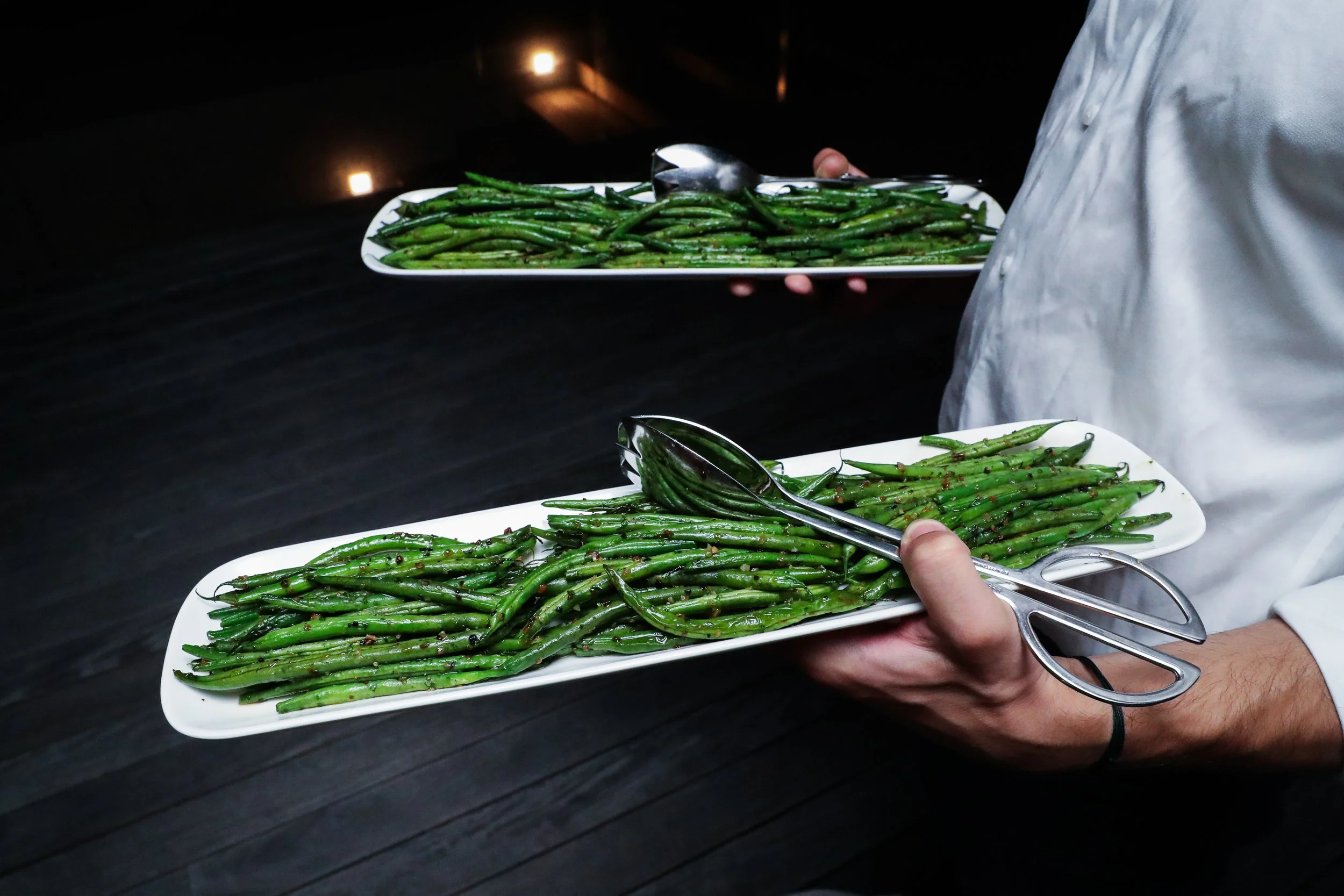 Two white rectangular plates filled with cooked green beans, being served by a person using silver tongs, against a dark background.