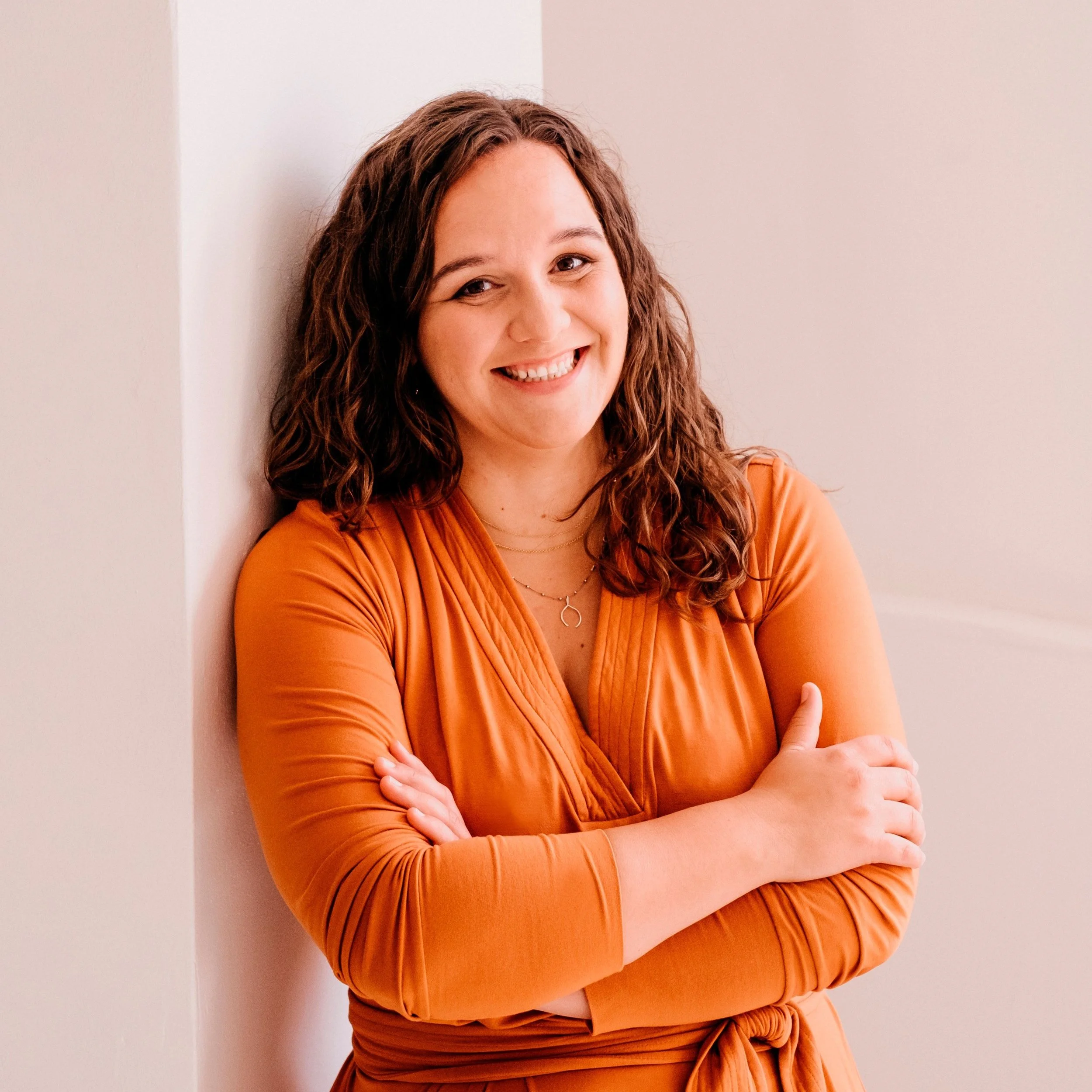 A woman with curly brown hair and a blue blouse sitting at a table with books, smiling and resting her chin on her hand.