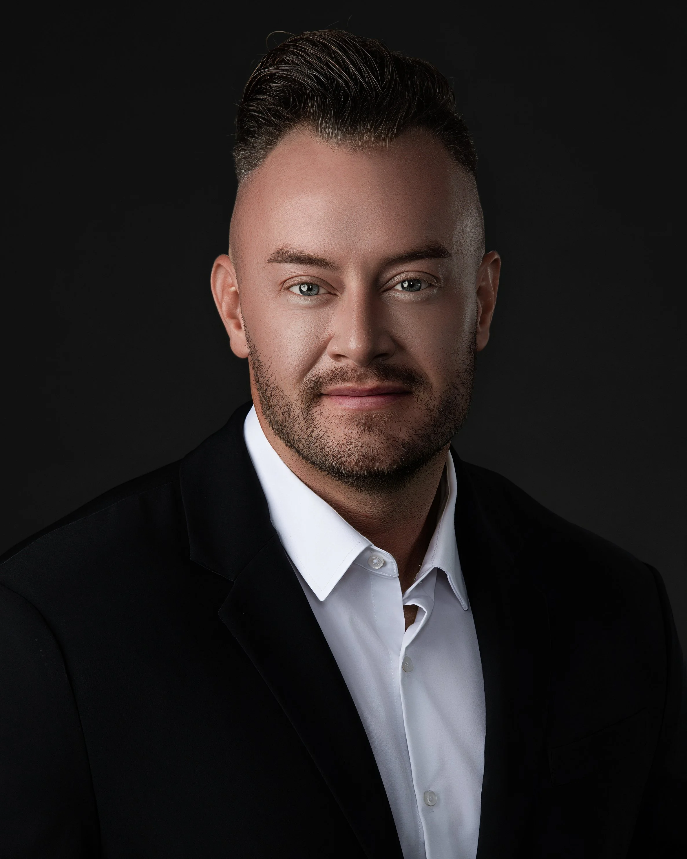 Close-up portrait of a man with short, styled dark hair, light skin, blue eyes, and a trimmed beard, wearing a black suit and white dress shirt, against a black background.
