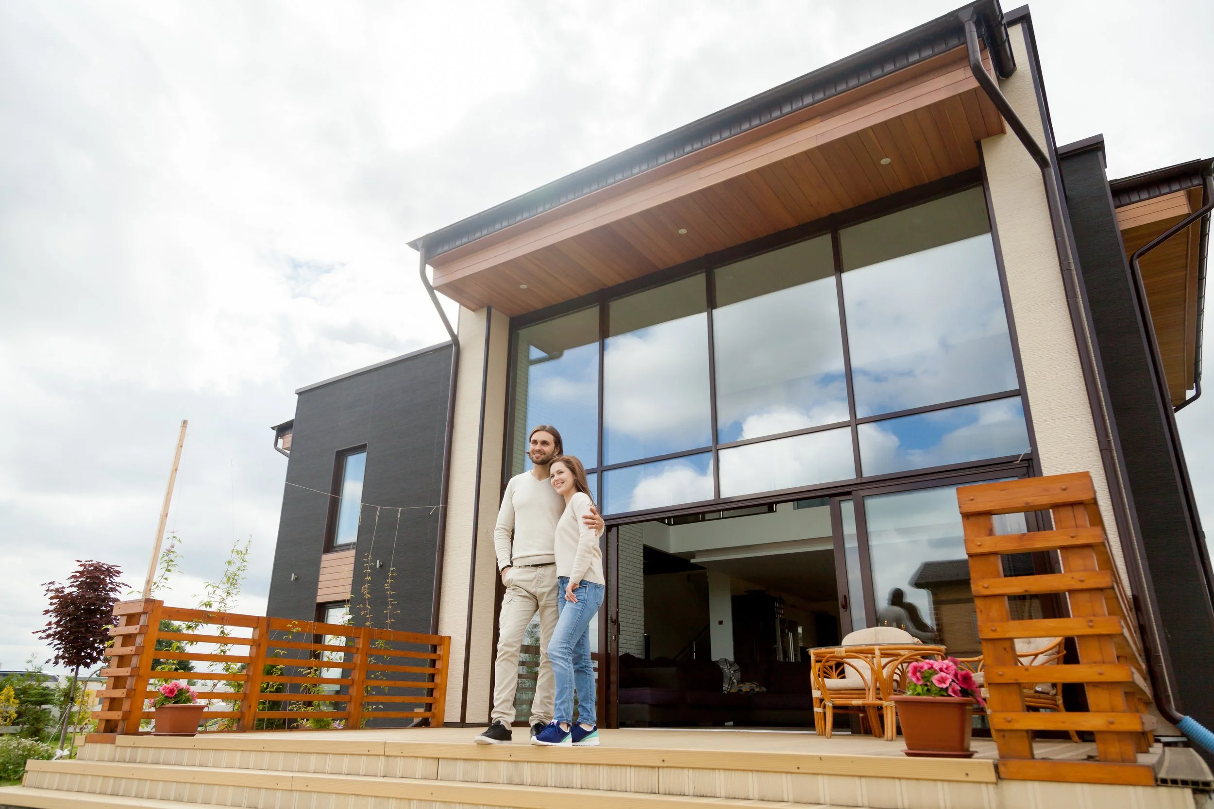 Two people, a man with long hair and a woman with shoulder-length hair, standing on a wooden deck outside a modern house with large glass windows and a sloped roof, under a cloudy sky, with some potted plants and outdoor furniture nearby.