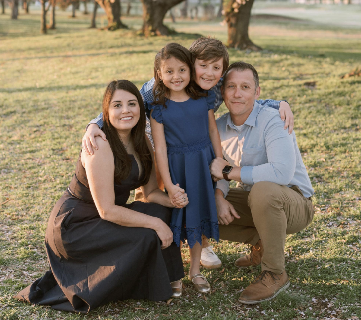 A family of four in a park during sunset, smiling and posing for a photo. The mother and father are kneeling, with their children standing between them. The girl is wearing a blue dress, and the boy is in a blue shirt. There are trees with pink blossoms in the background.
