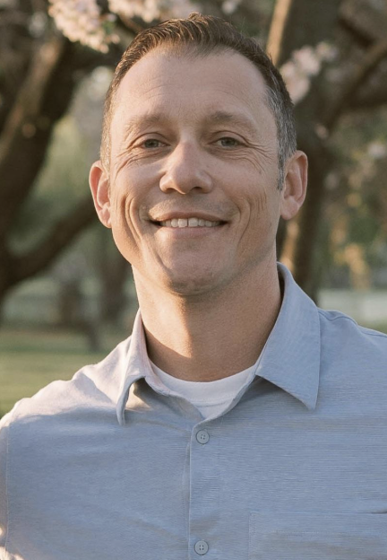 A smiling man wearing a light blue collared shirt outdoors with trees and sunlight in the background.