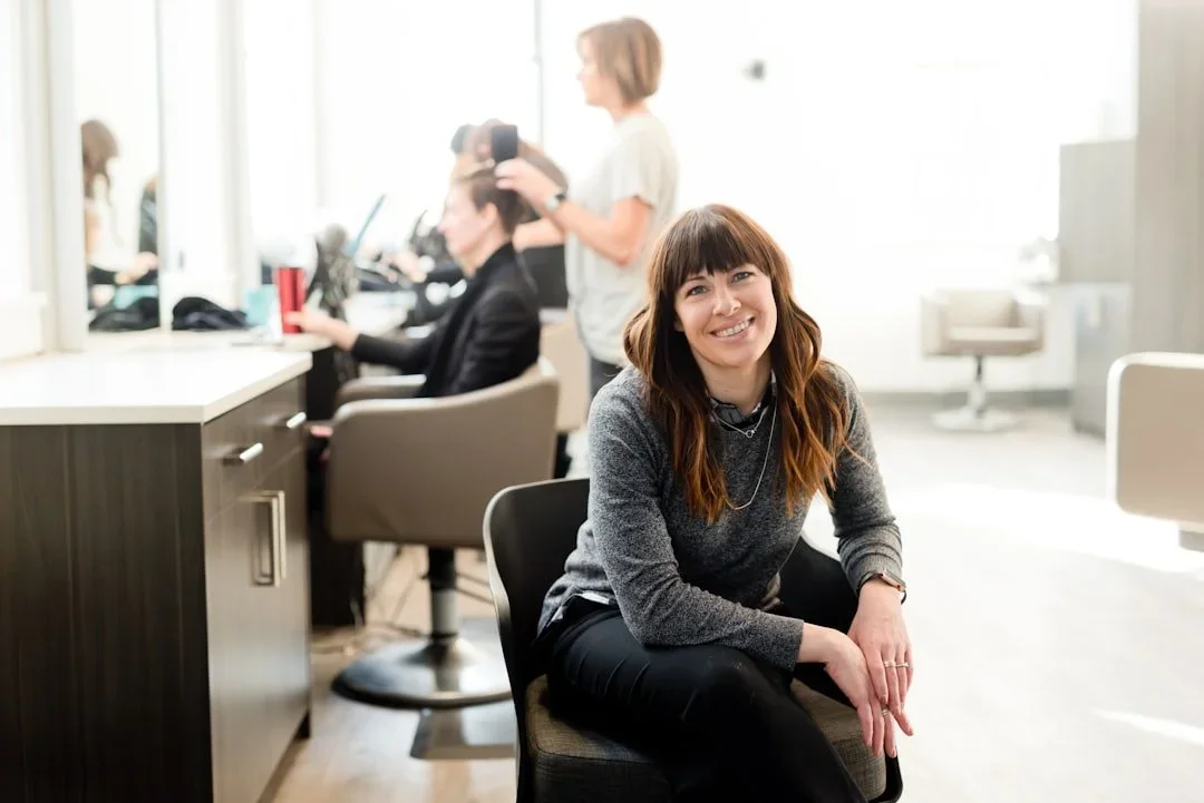 A woman with long brown hair and a gray sweater smiling while sitting in an office, with colleagues working in the background.