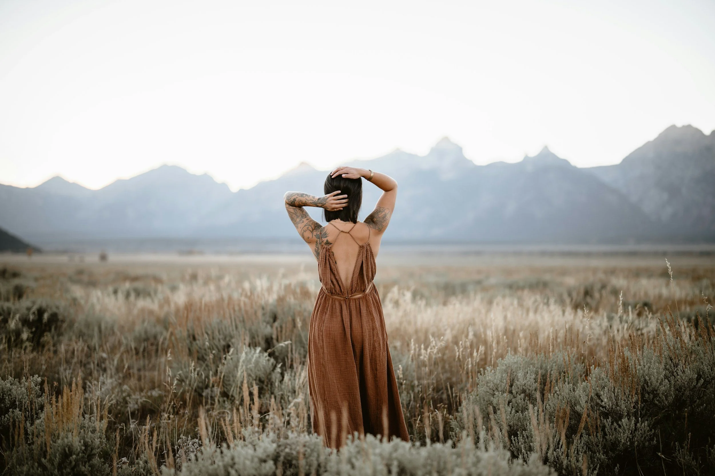 A woman with tattoos on her arms wearing a brown dress stands in a field with mountains in the background, looking at the mountains with her hands on her head.