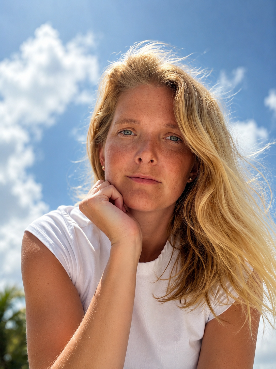A woman with blonde hair smiling, wearing sunglasses and a light-colored shirt, sitting near a dock with boats in the background.