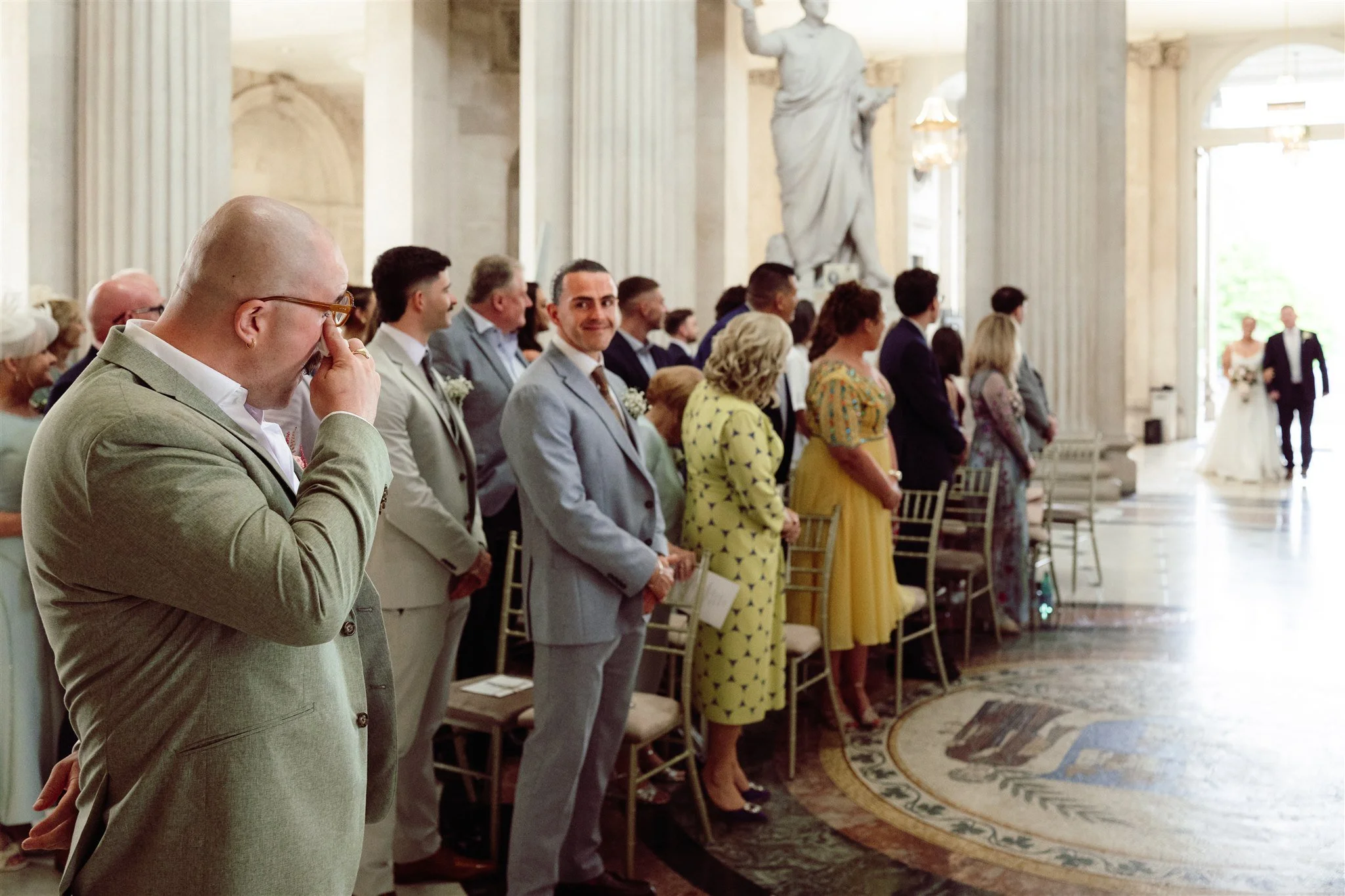 People attending a wedding ceremony inside a grand hall with tall columns and statues. The groom and bride are visible in the background walking down the aisle.