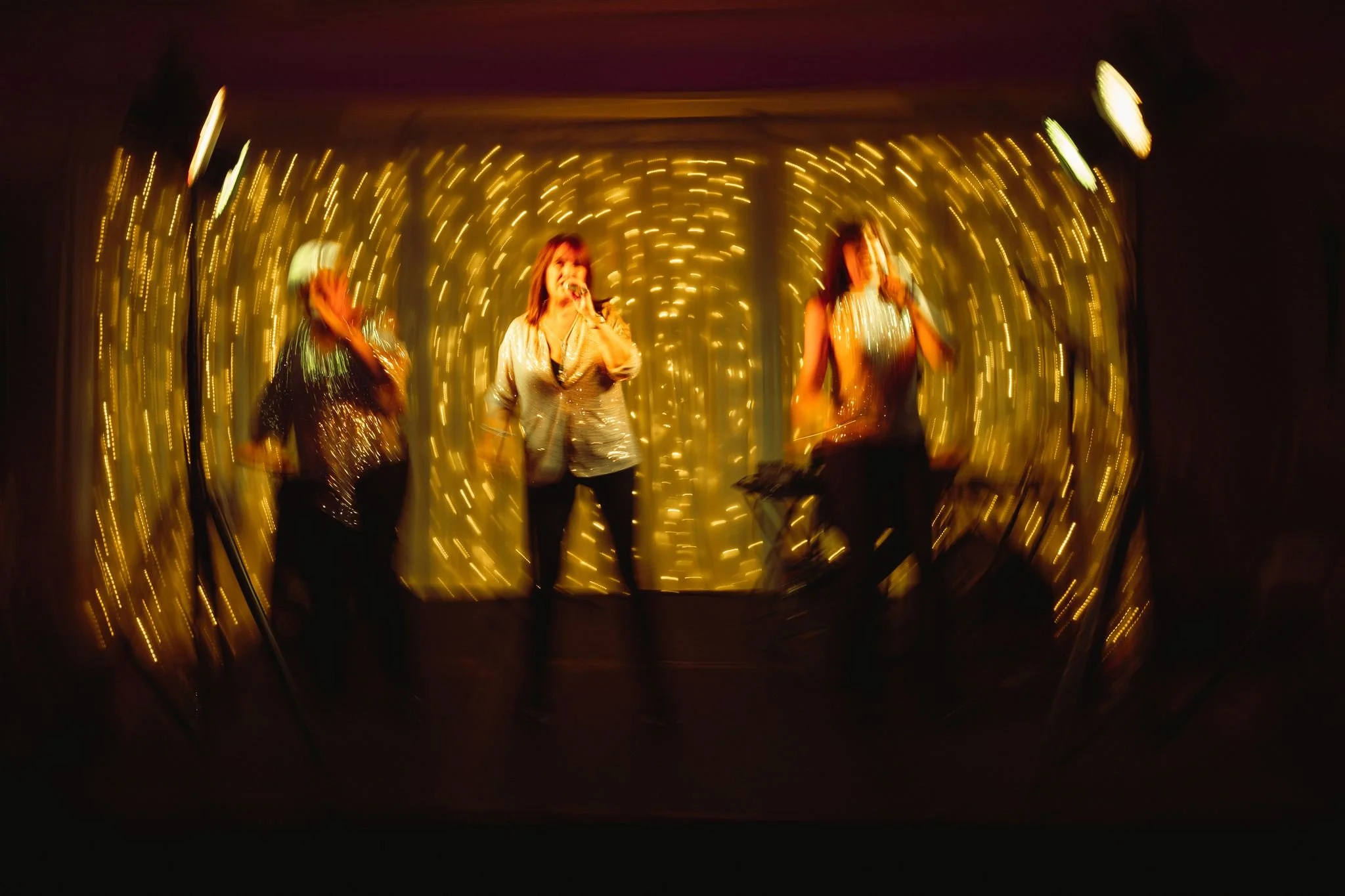 Three women singing and playing music on a stage decorated with string lights, with a blurred, swirling effect.