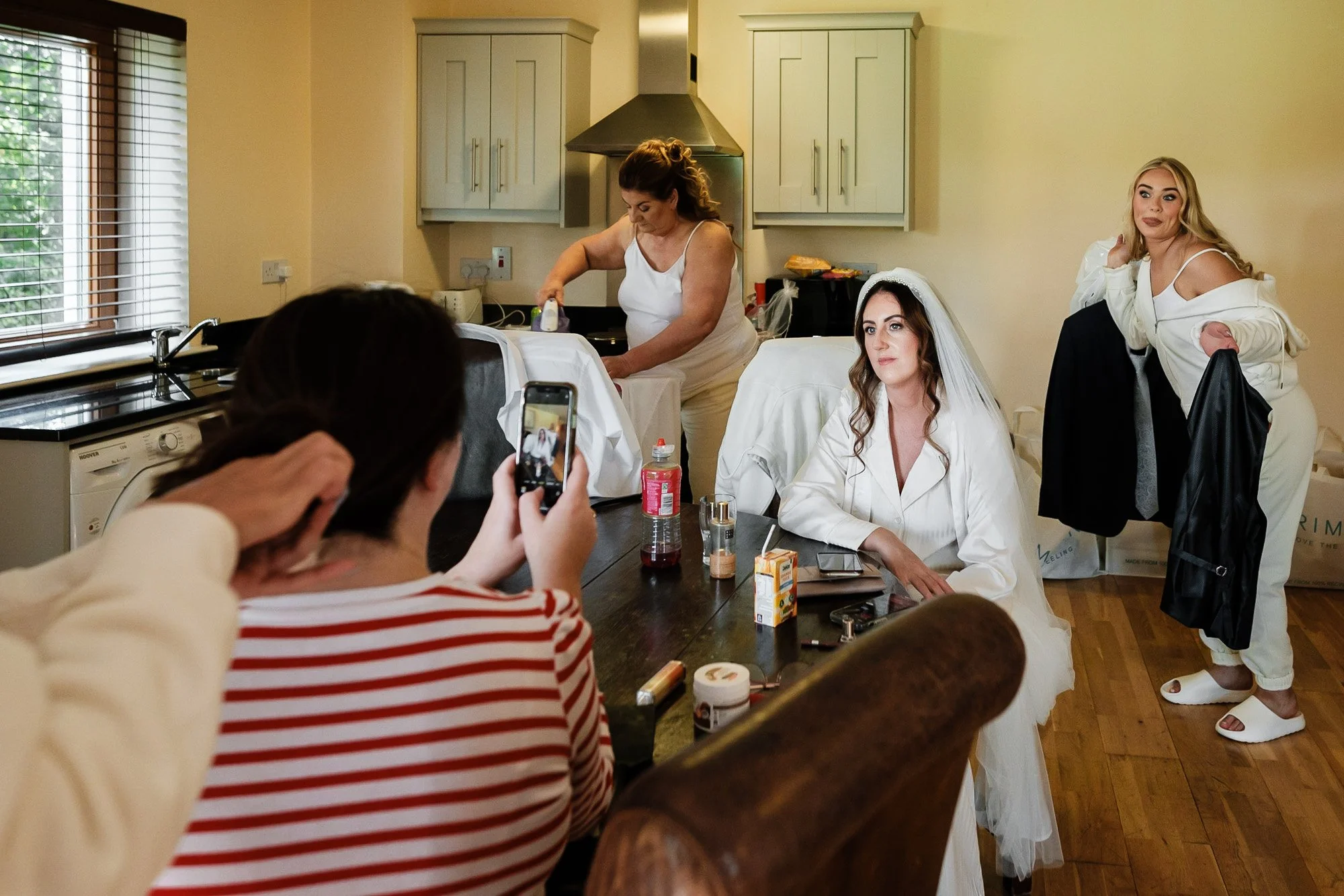 Women preparing for a wedding in a kitchen, one woman is ironing a white garment, another is sitting at a table with makeup and beauty products, and a third woman is sitting wearing a veil, while another woman stands nearby holding a garment bag.