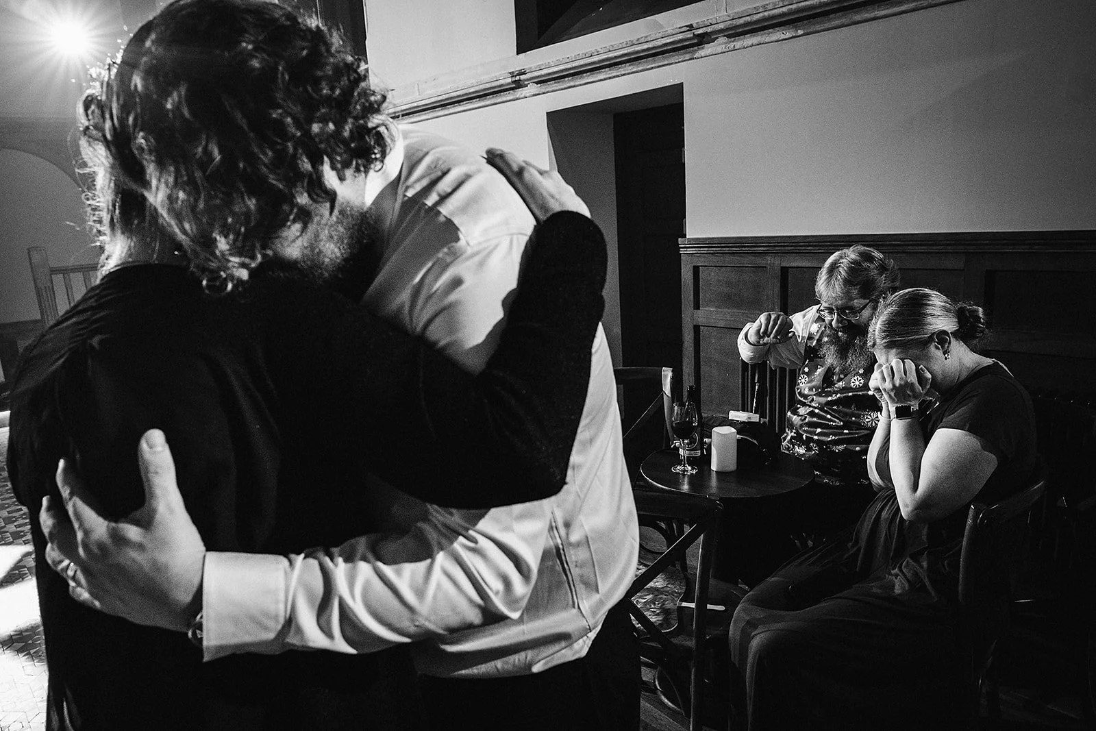 A couple is dancing closely, embracing each other, while two women sit nearby, one with her head down and the other adjusting her glasses with her head bowed, in a dimly lit room with wine glasses on a table.