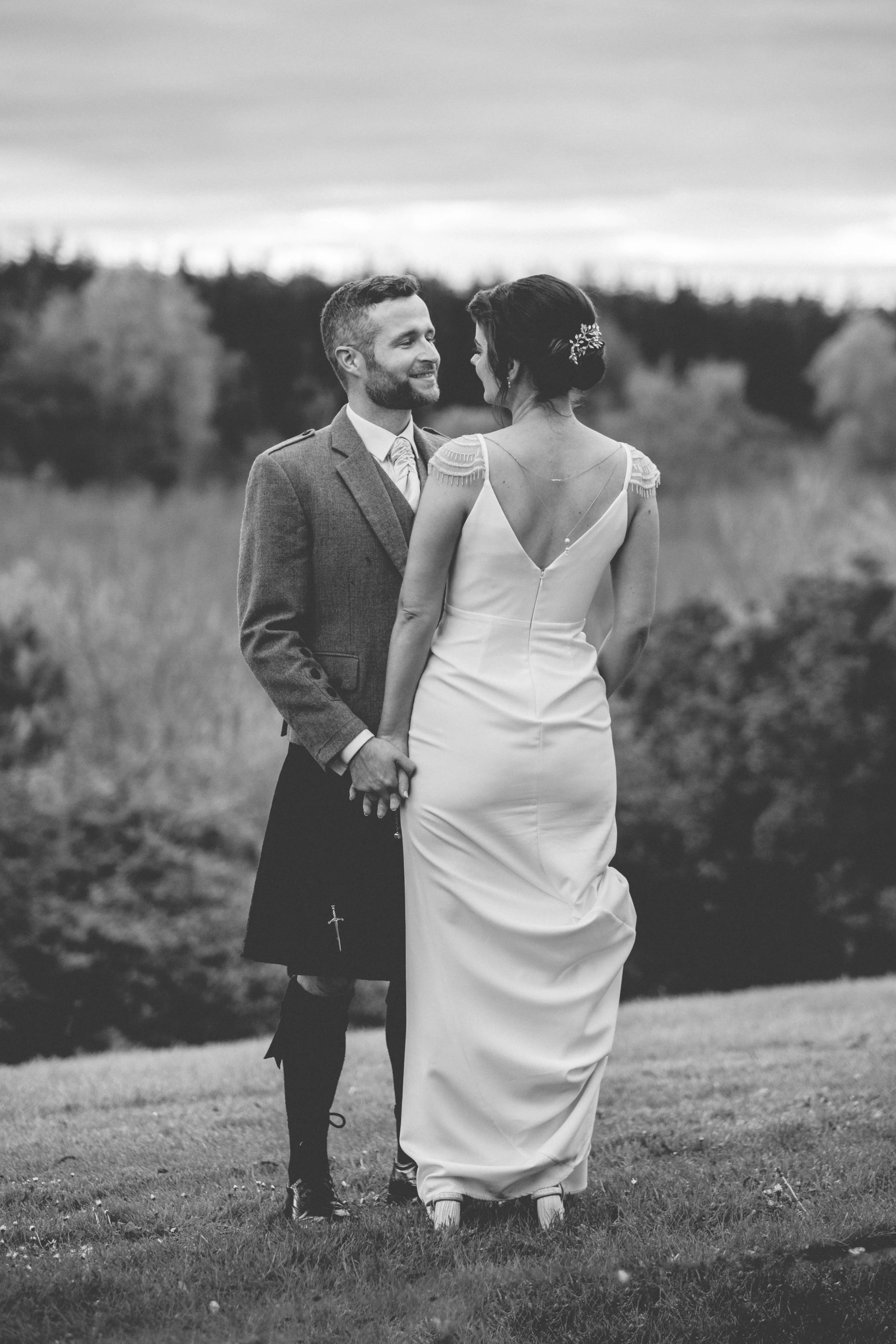 A black and white photograph of a couple in wedding attire standing outdoors on a grassy field, holding hands and looking at each other with smiling faces, with a background of trees and a cloudy sky.