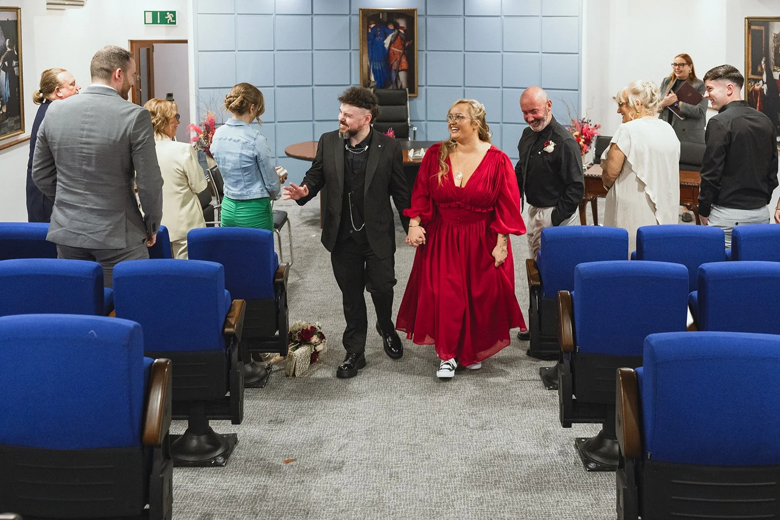People gathering in a room for a celebration, with a woman in a bright red dress holding hands with a man in black, walking down the aisle as others smile and watch, some standing and some seated.