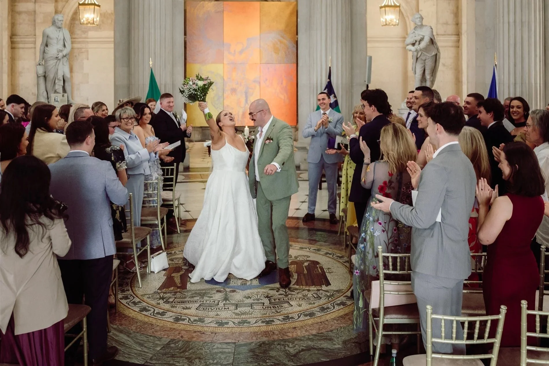 Bride and groom smiling and holding a bouquet of flowers in the middle of a wedding ceremony inside a grand hall with guests clapping and celebrating.