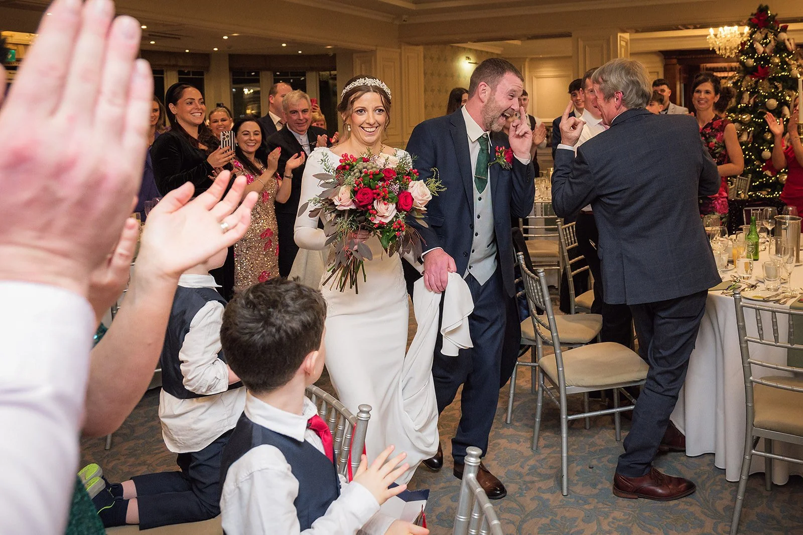 Bride and groom walking down the aisle at their wedding reception, surrounded by guests clapping and celebrating, with a decorated Christmas tree in the background.
