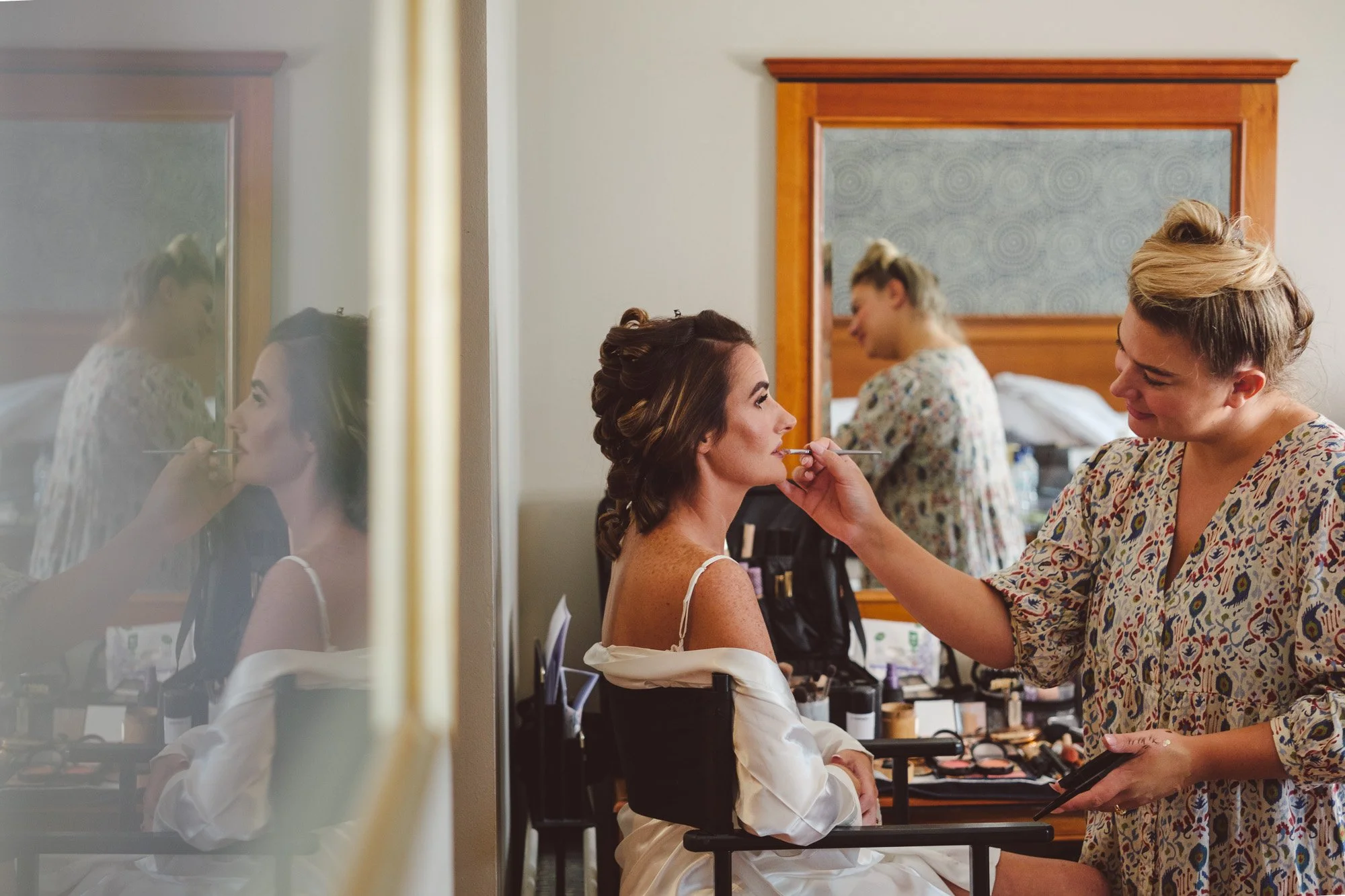 Bridal being prepared by a makeup artist in a hotel room, with a friend in the background.